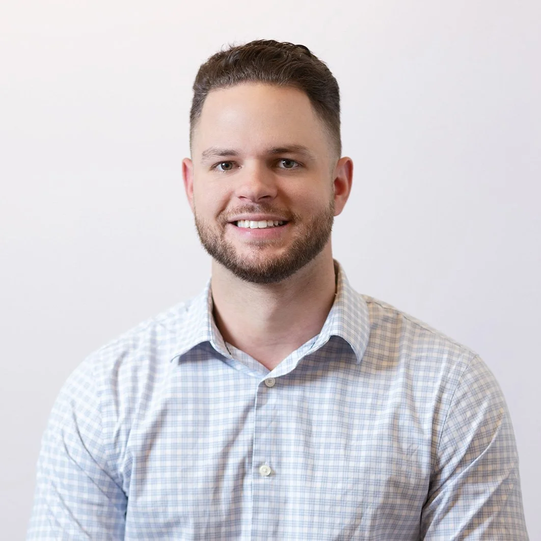 Headshot of a smiling man with short brown hair and beard, wearing a light plaid shirt, against a plain white background.