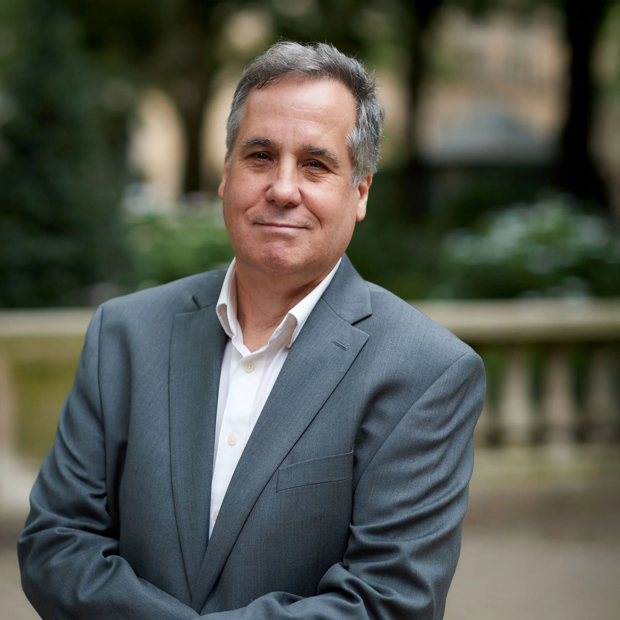 Portrait of a mature man in a gray suit and white shirt, standing outdoors with a blurred natural background.