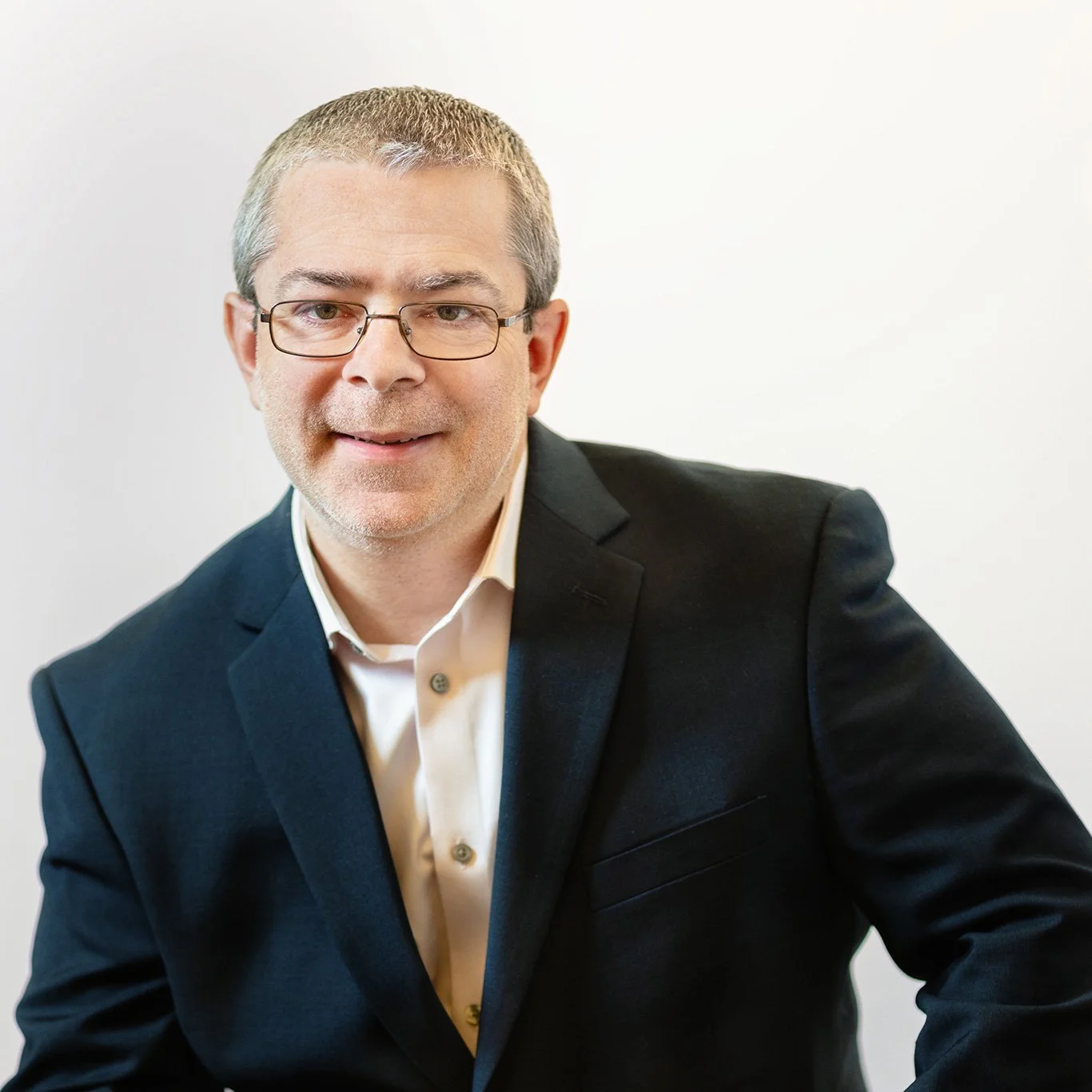 A middle-aged man in a dark suit jacket, white shirt, wearing glasses, smiling, against a plain white background.