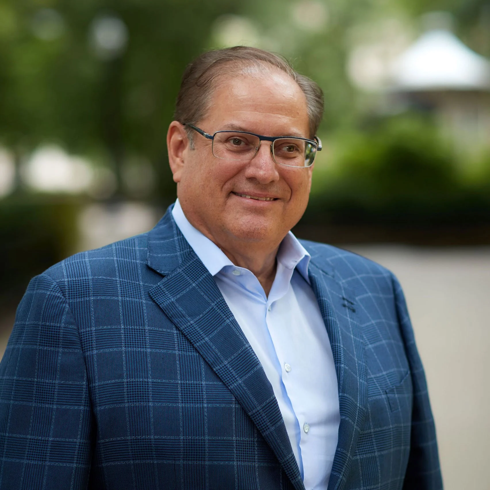 A middle-aged man wearing glasses and a blue checkered suit smiling outdoors with trees in the background.