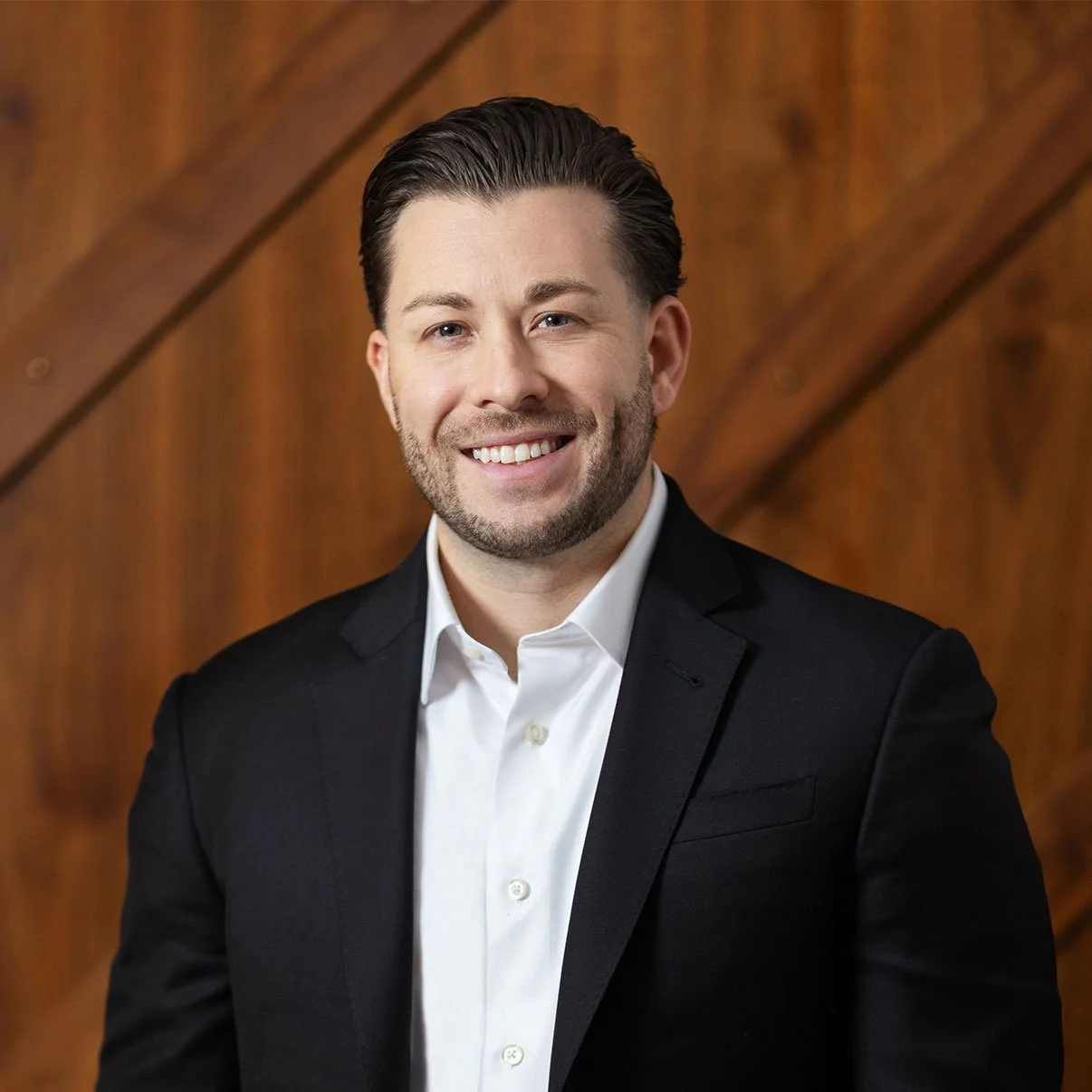 A smiling man with dark hair and stubble, dressed in a black suit and white shirt, standing in front of a wooden background.