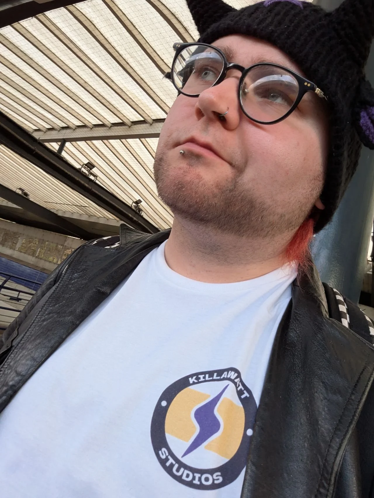 A person with glasses, piercings, and a black beanie looking upwards. They are wearing a white t-shirt with a logo and a leather jacket, and are at an outdoor location with a structure overhead.