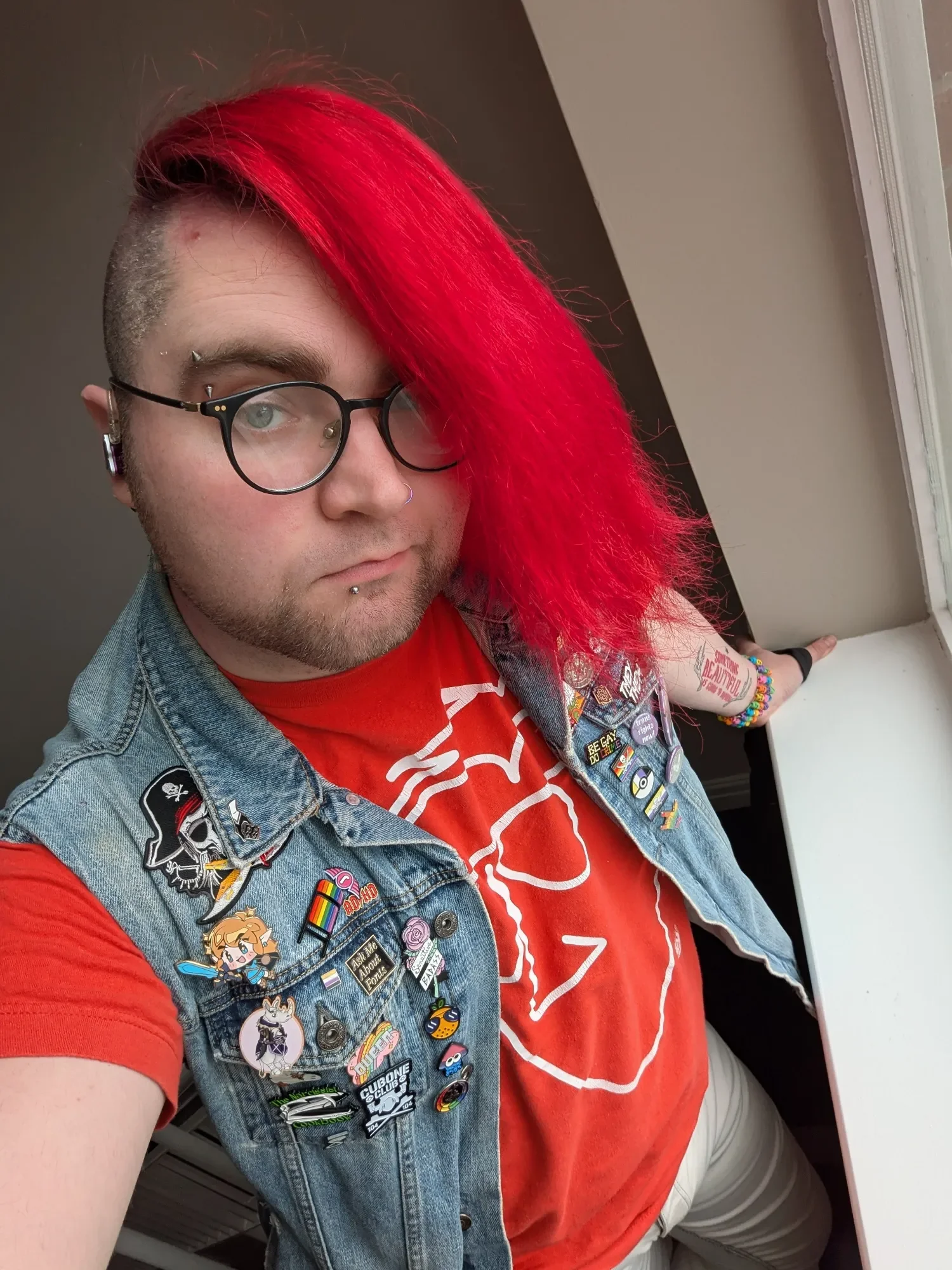 A person with bright red hair, wearing glasses, a denim vest with numerous pins and patches, and a red t-shirt with a white outline of an outline of an animal, standing near a window.