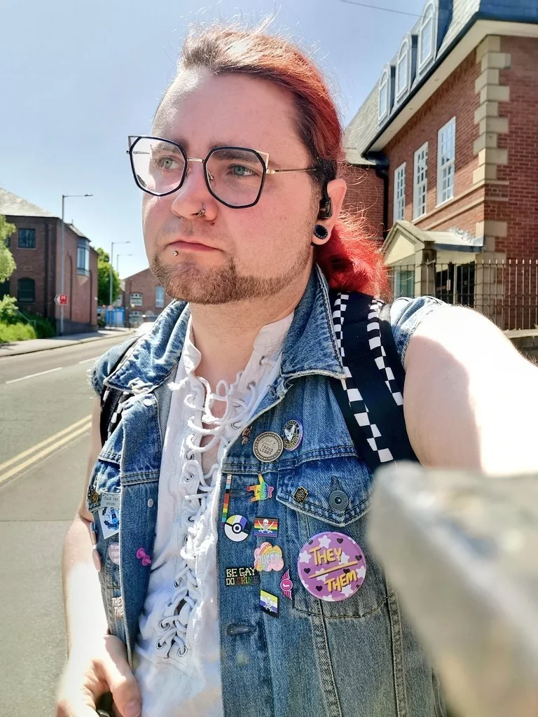 Person with red hair, glasses, and facial piercings taking a selfie outdoors in front of a building on a sunny day, wearing a denim vest with various pins and badges.