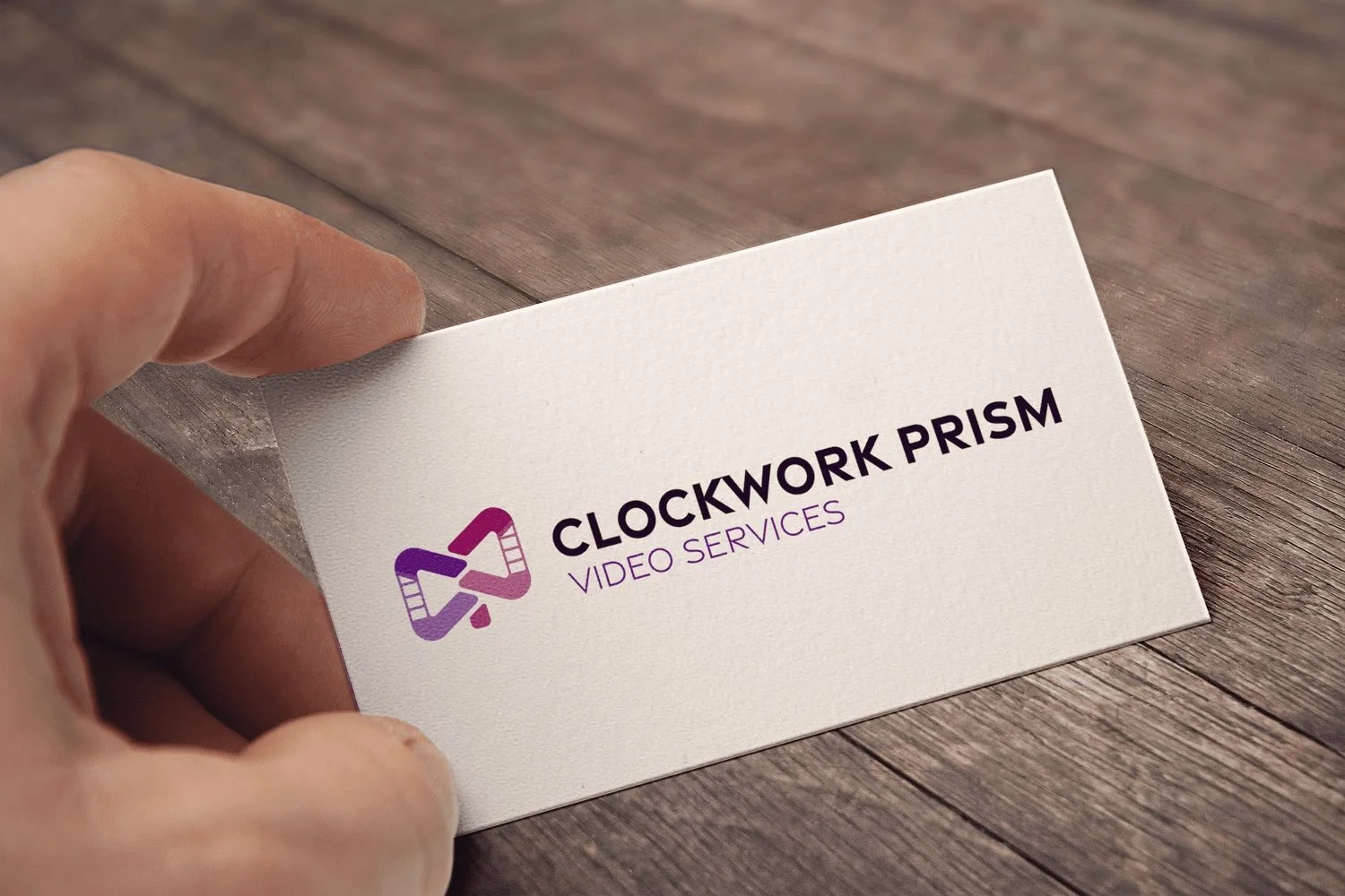 Business card for Clockwork Prism Video Services, featuring purple and black logo, held by a person's hand on a wooden surface.