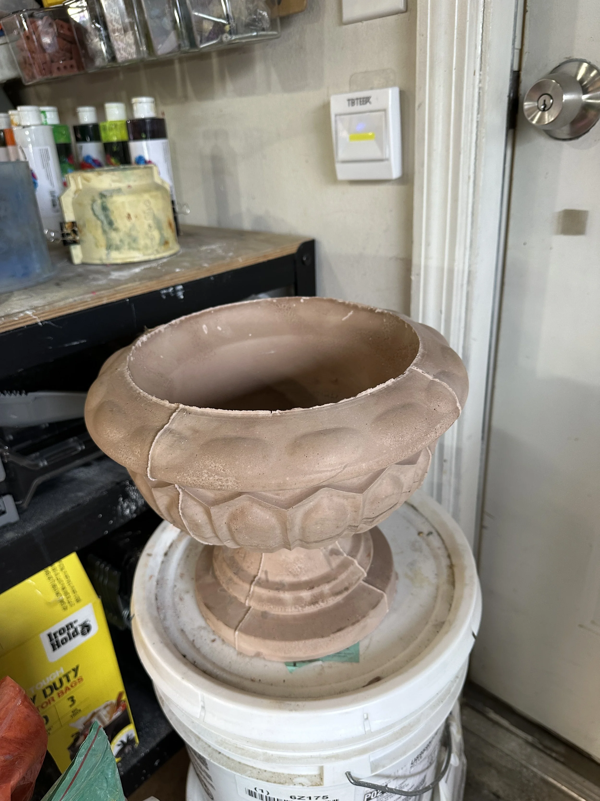 A large, unfinished, decorative flower pot with a scalloped edge, resting on a white plastic bucket in a workshop or studio.