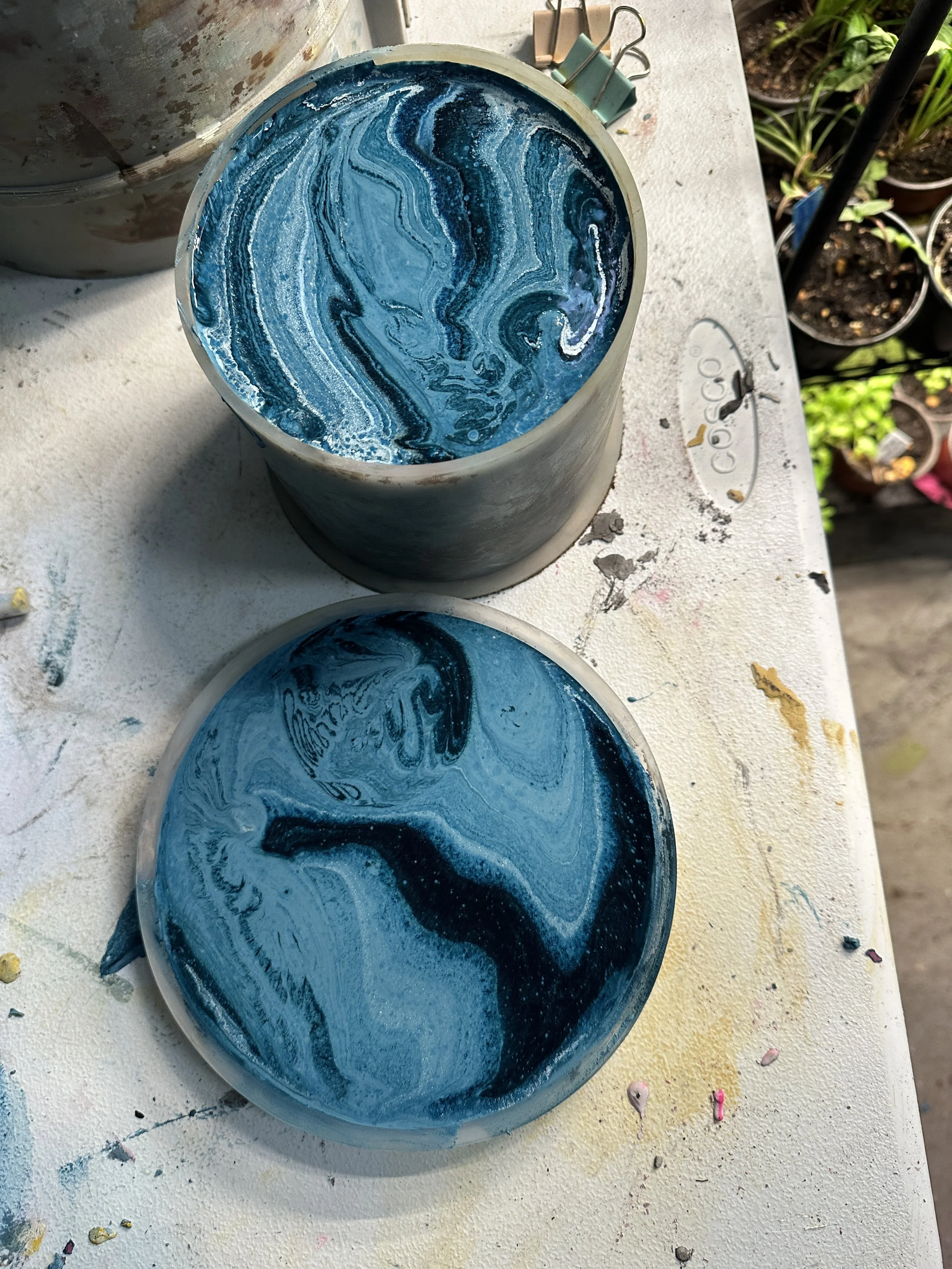 Blue and white marbled cement in a mold on a paint-stained work surface. A lid with matching marbled pattern is placed nearby. Gardening pots are visible in the background.