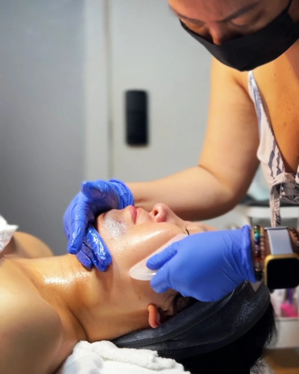 A woman lying down with her face relaxed as a person in blue gloves applies a facial treatment, with a mask or cream being used in a skincare procedure.