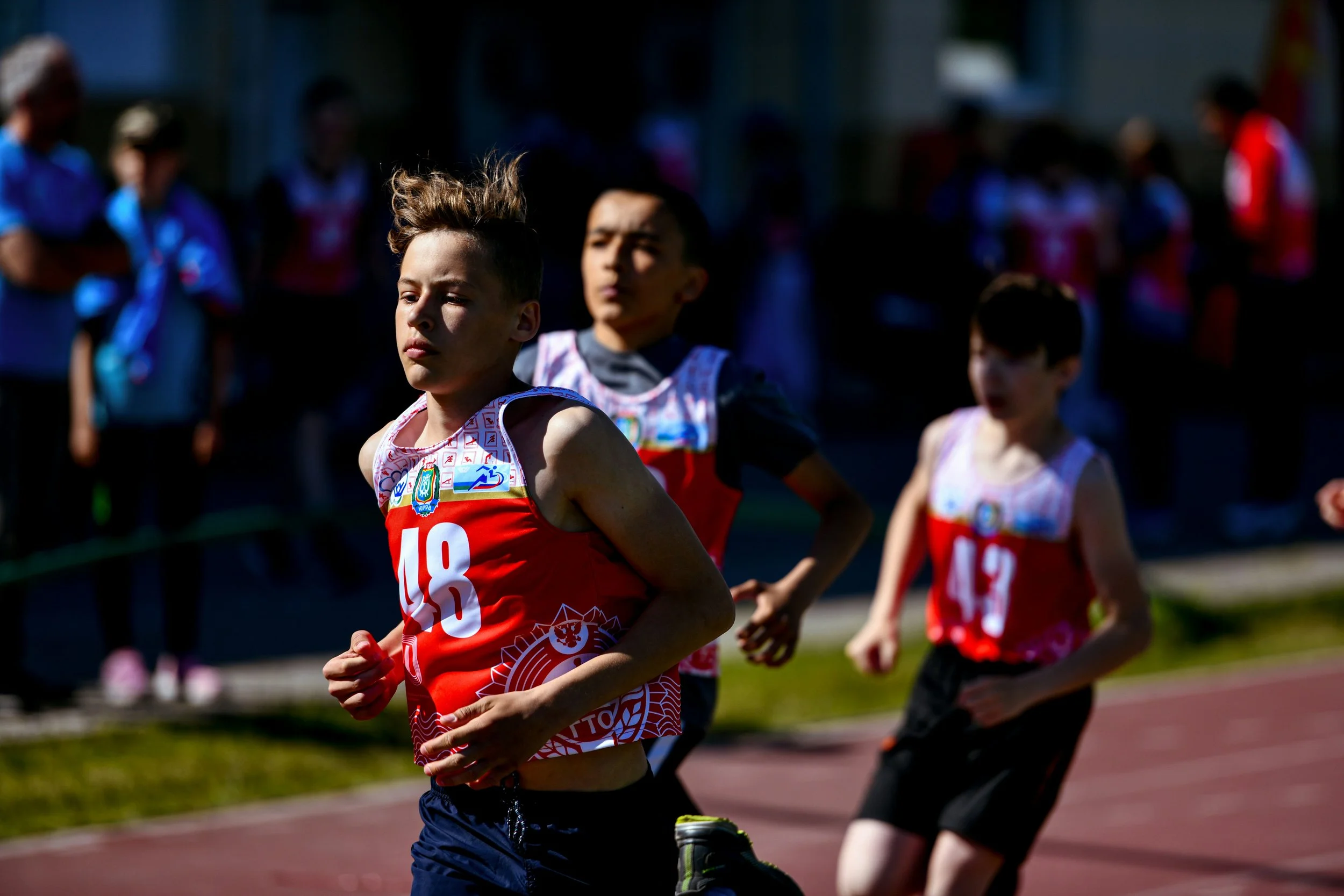 Children running in a race on a track, wearing athletic uniforms with numbers, with spectators watching in the background.