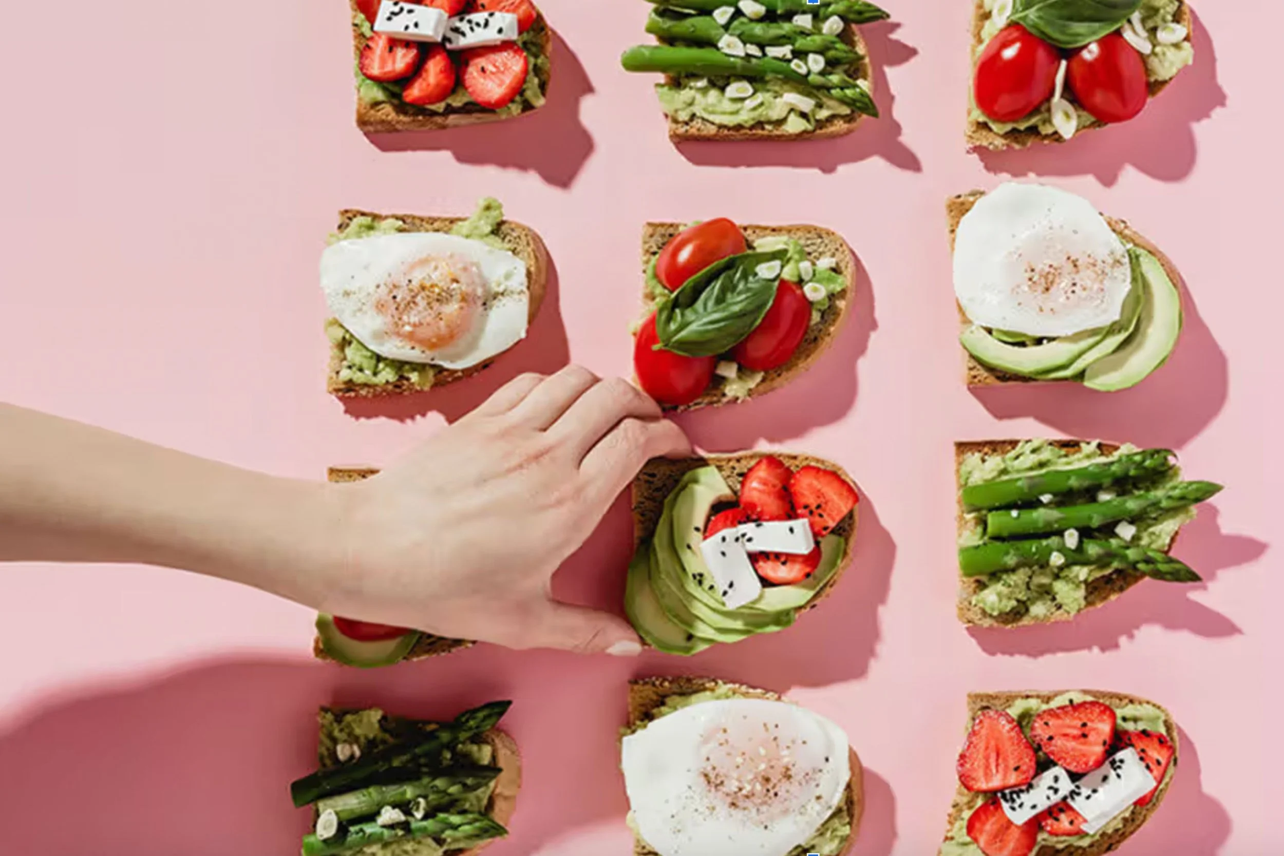 A hand arranging small open-faced sandwiches on a pink surface. The sandwiches are topped with various ingredients including strawberries, cherry tomatoes, asparagus, avocado, eggs, basil, and dragon fruit, each on slices of bread or toast.