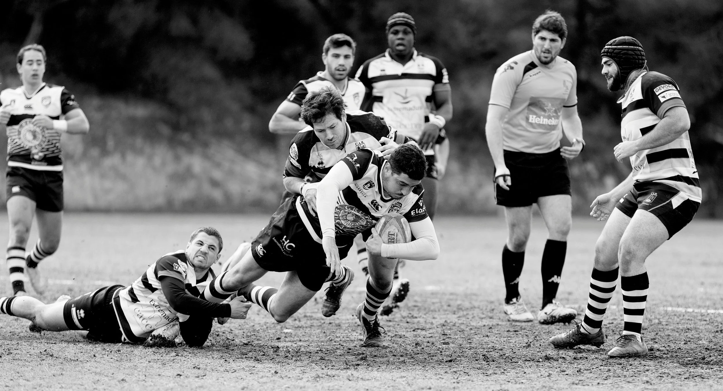A rugby match with players in action, one player running with the ball while others attempt to tackle him, on a grassy field, black-and-white photo.
