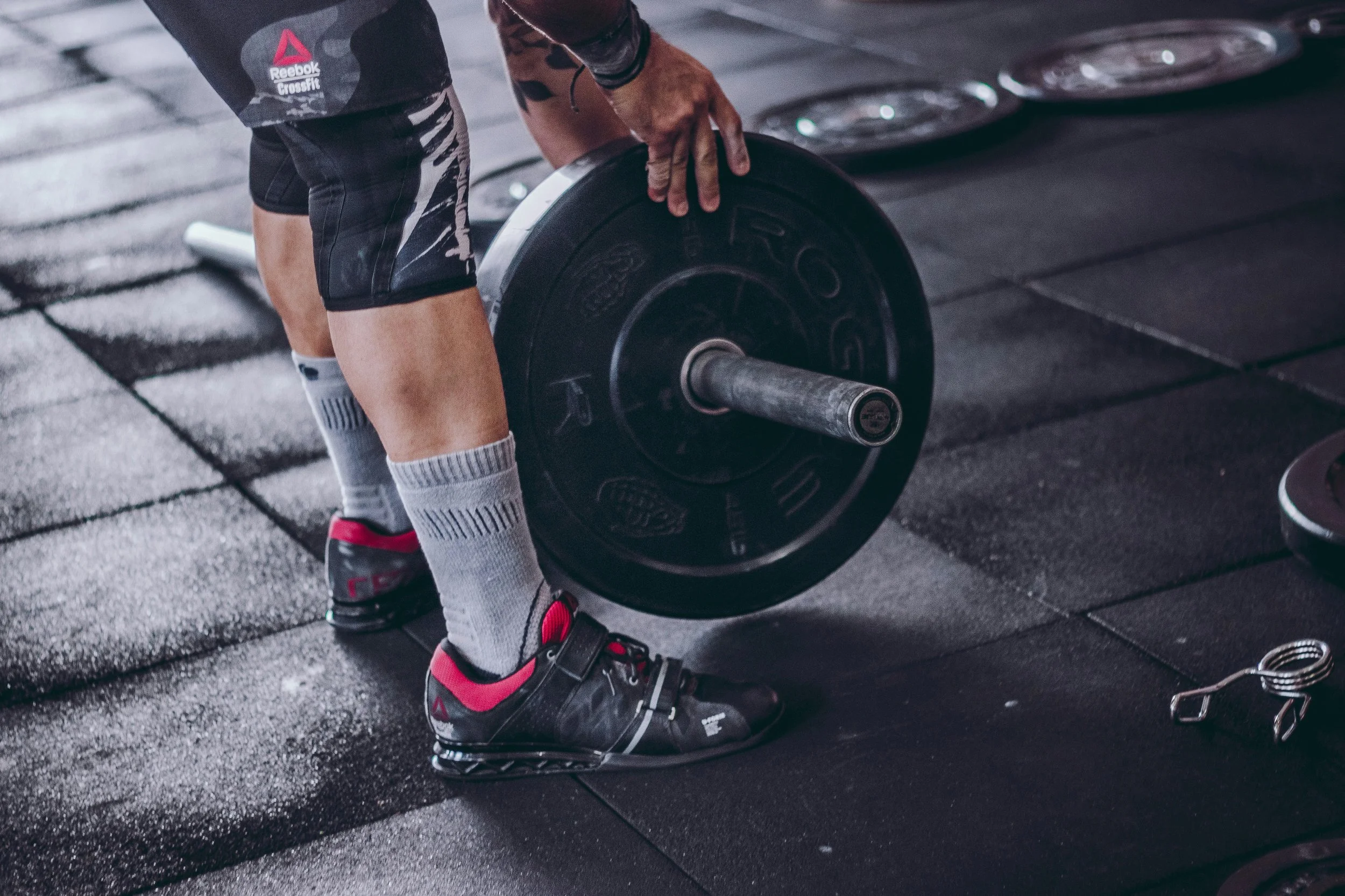 Person wearing black and red athletic shoes, gray socks, and black shorts lifting a barbell with weight plates in a gym.