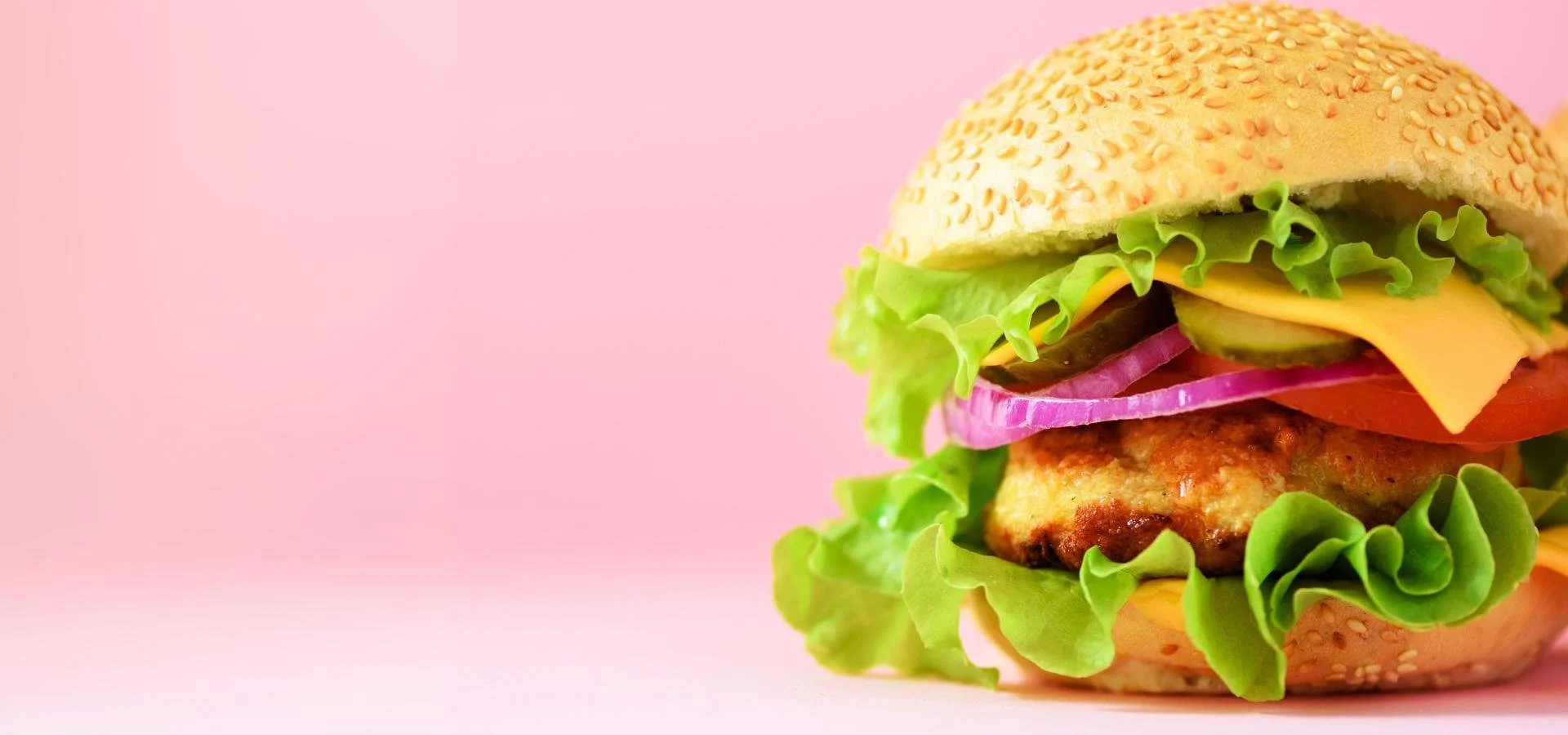Close-up of a burger with sesame seed bun, lettuce, cheese, tomato, pickles, red onions, and a grilled chicken patty against a pink background.