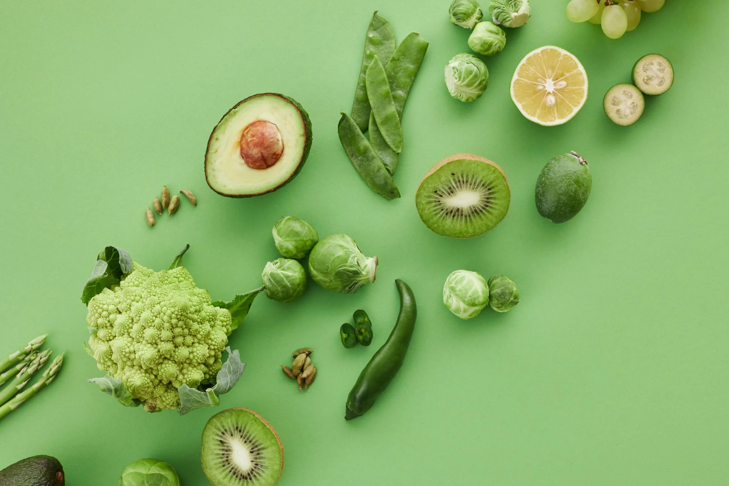 Various green fruits and vegetables on a light green background, including an avocado, Brussels sprouts, kiwi, lemon, cucumber, Brussels sprout leaves, green grapes, scallions, a lime, and sunflower seeds.