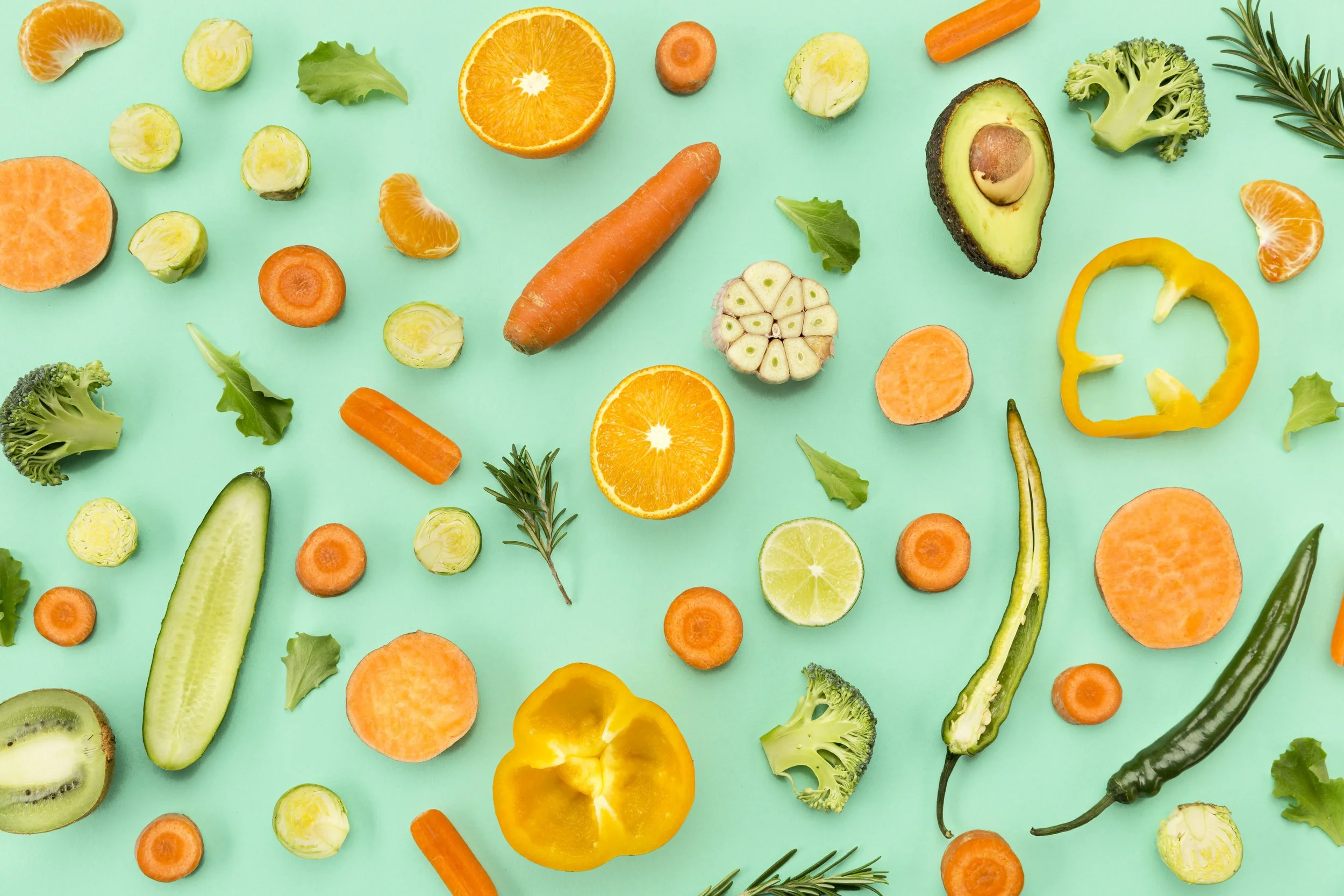 A flat lay of various cut and whole fresh vegetables and fruits, including carrots, bell peppers, broccoli, cucumber, avocado, citrus fruits, garlic, and herbs on a light green background.