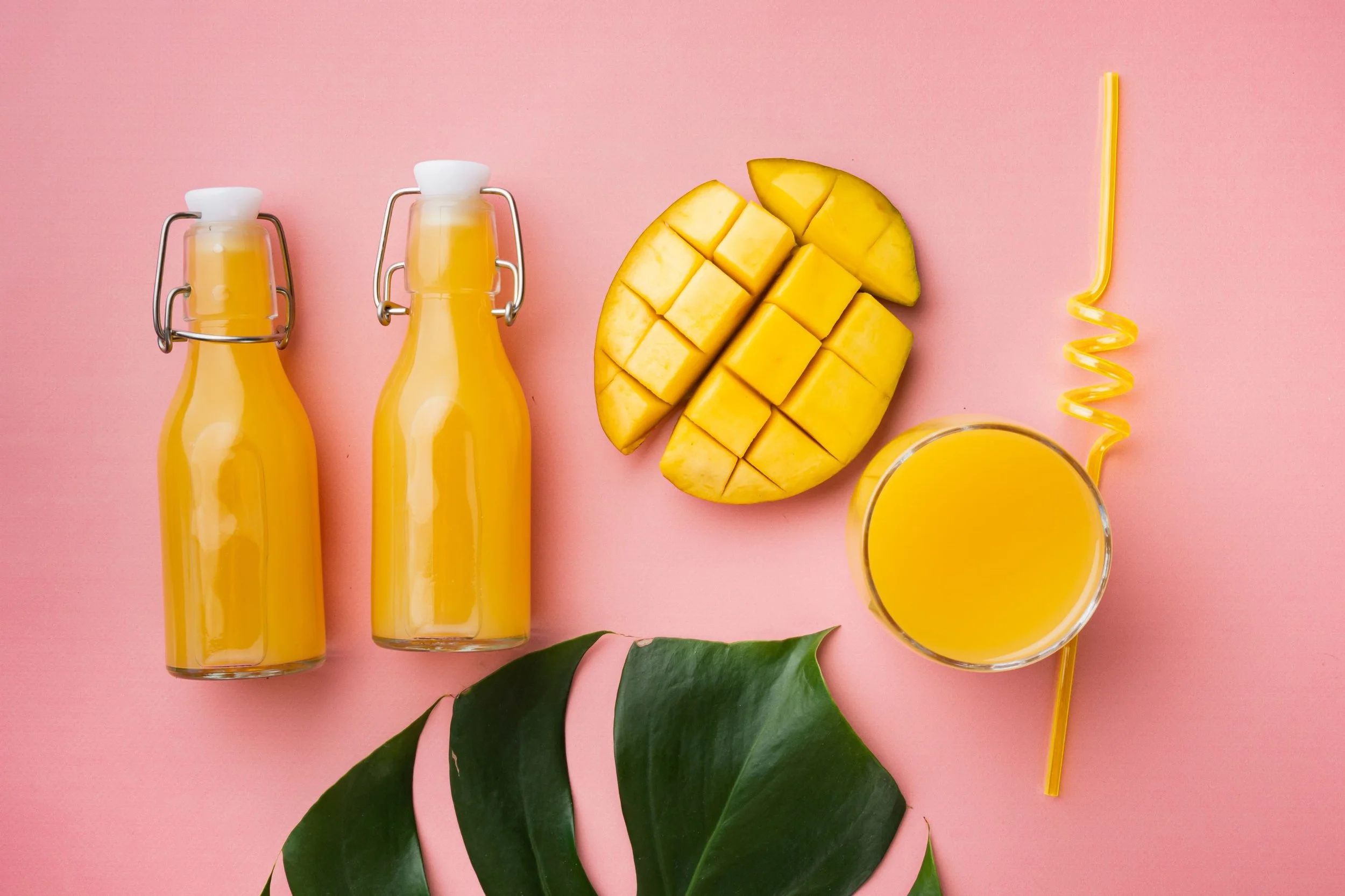Two bottles of mango juice, a sliced mango, a glass of mango juice, and a yellow curly straw on a pink background with green leaves.