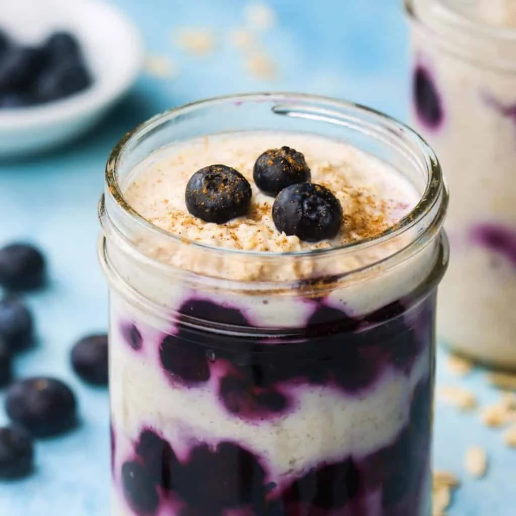 A glass jar filled with a layered blueberry yogurt parfait, topped with three blueberries and sprinkled with cinnamon or nutmeg, with blueberries scattered nearby.