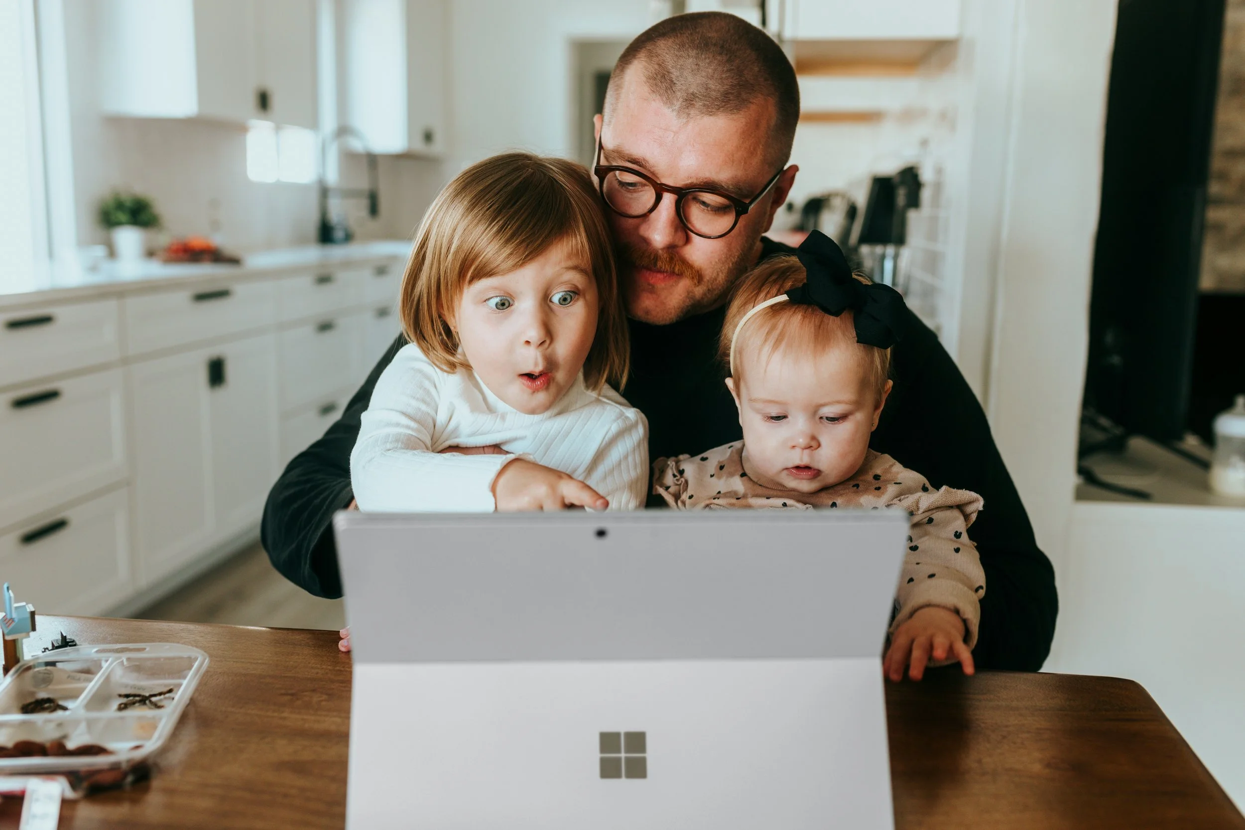 A man with glasses and a beard, two young girls, one with a surprise expression, are sitting at a wooden table looking at a laptop in a bright, modern kitchen.