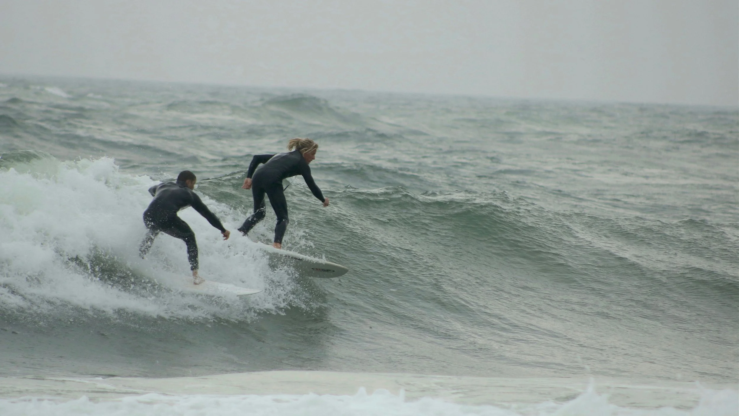 Two people surfing on a wave in the ocean, both wearing wetsuits.