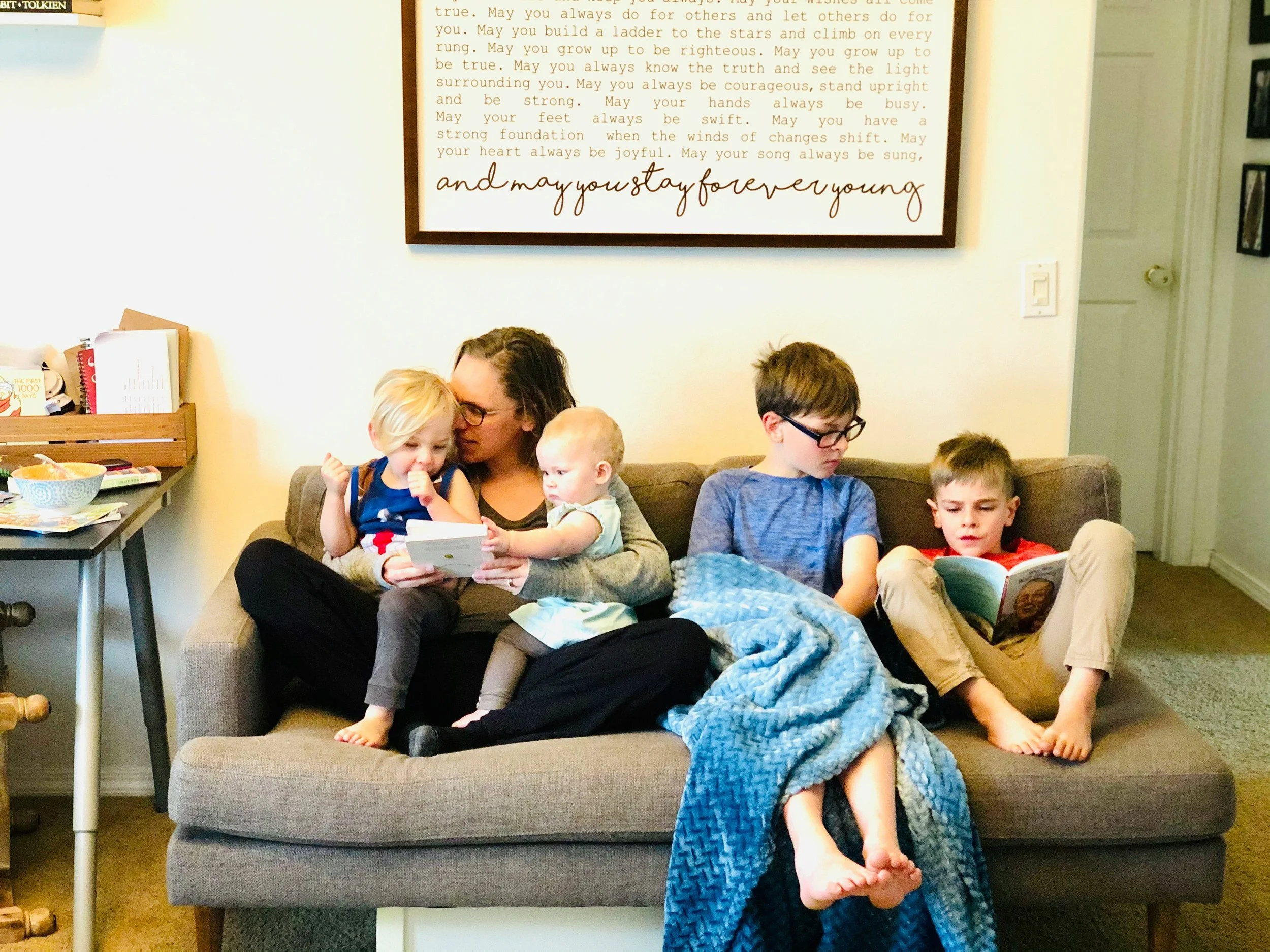 A woman sitting on a beige sofa with four children, each engaged in reading or looking at a book, in a cozy living room. A blanket covers some of the children. There is a wall art behind them with a quote about staying young and true. To the left, a small table with books and snacks.