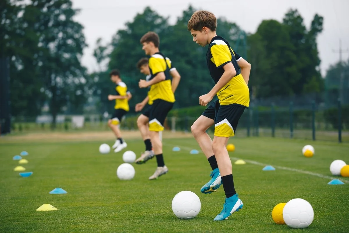 Young boys in yellow and black sports uniforms practicing soccer. They are kicking soccer balls on a field with cones for drills, with trees in the background.
