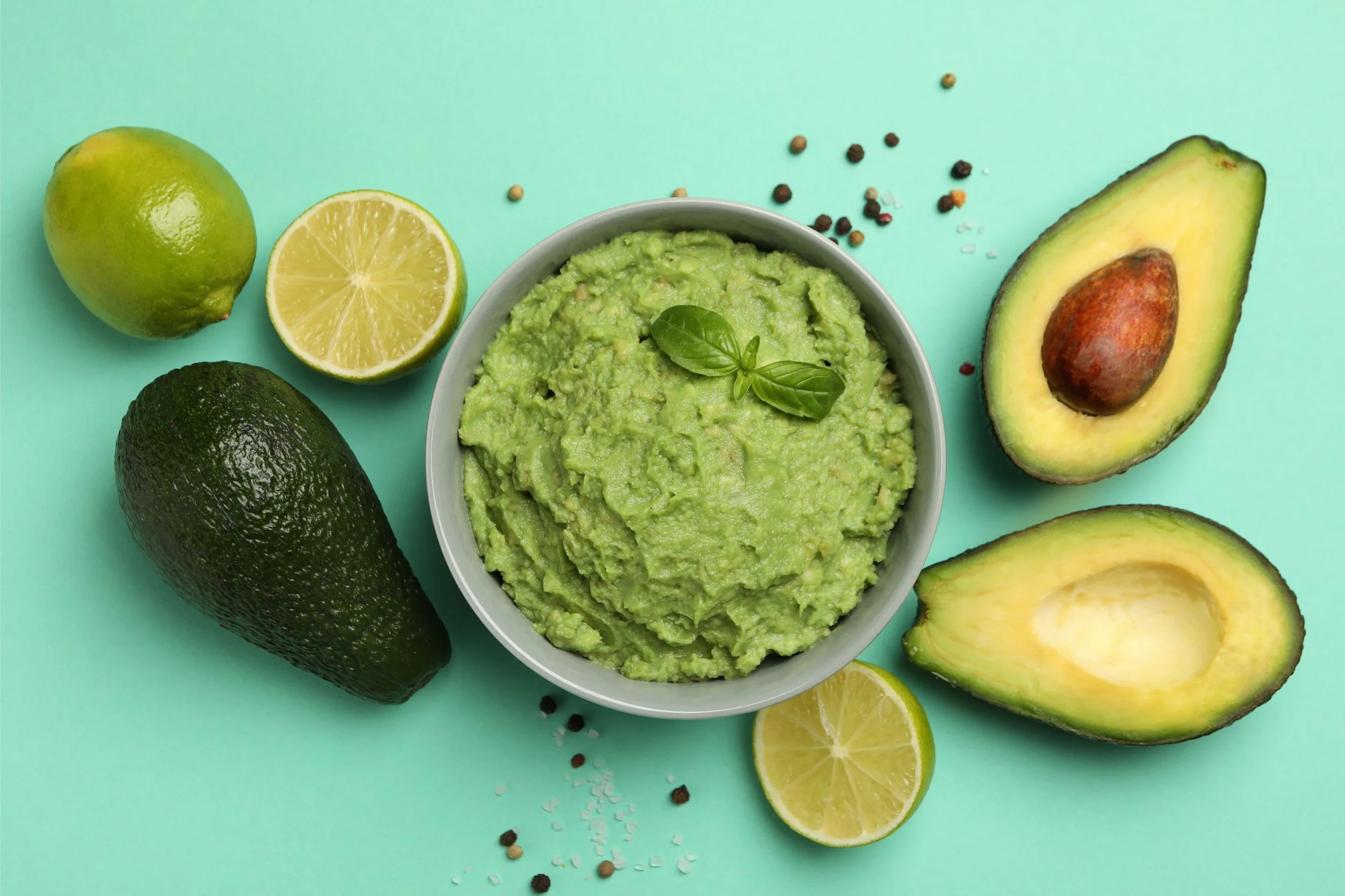 A bowl of guacamole garnished with a basil leaf surrounded by whole and halved avocados, limes, and scattered peppercorns on a turquoise background.