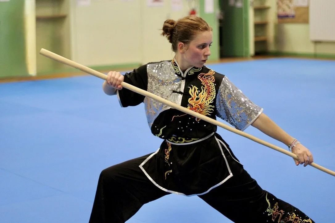 Jeune femme en costume traditionnel chinois pratiquant le kung-fu avec un bâton dans un gymnase.