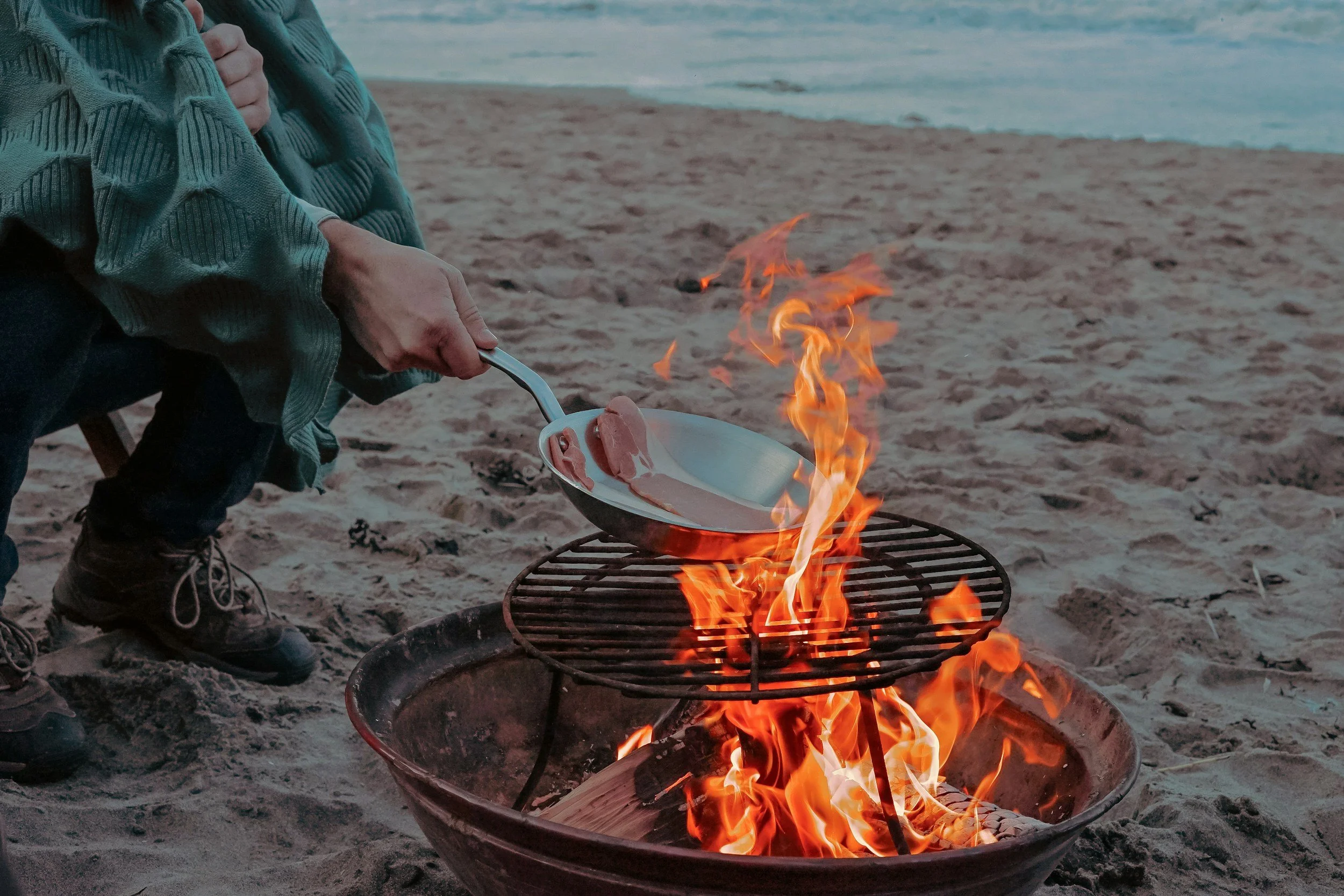 Person in a striped sweater roasting slices of ham over an open fire on a beach with the ocean in the background.