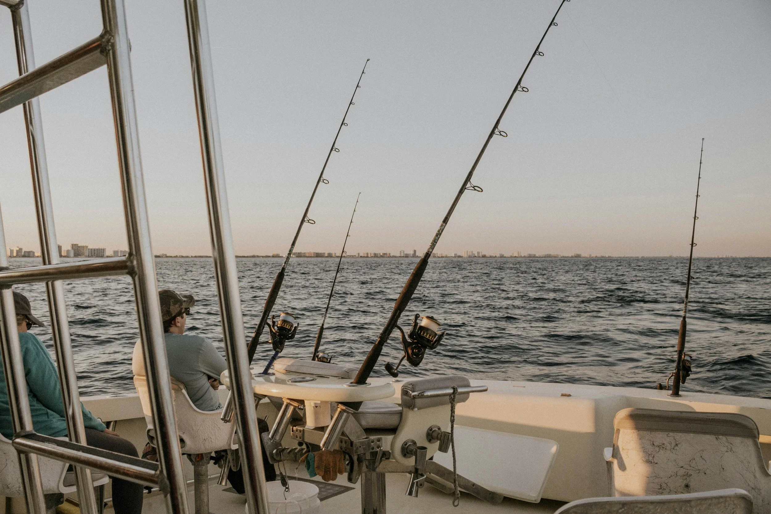People sitting on a boat fishing in the water during sunset with a city skyline in the distance.