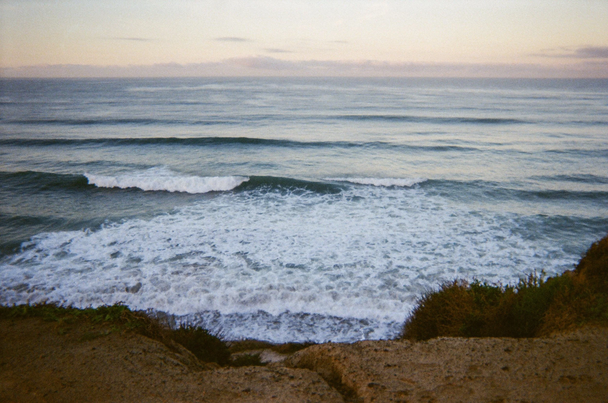 Photo of ocean waves crashing on the shore with rocks and greenery in the foreground, under a clear sky.