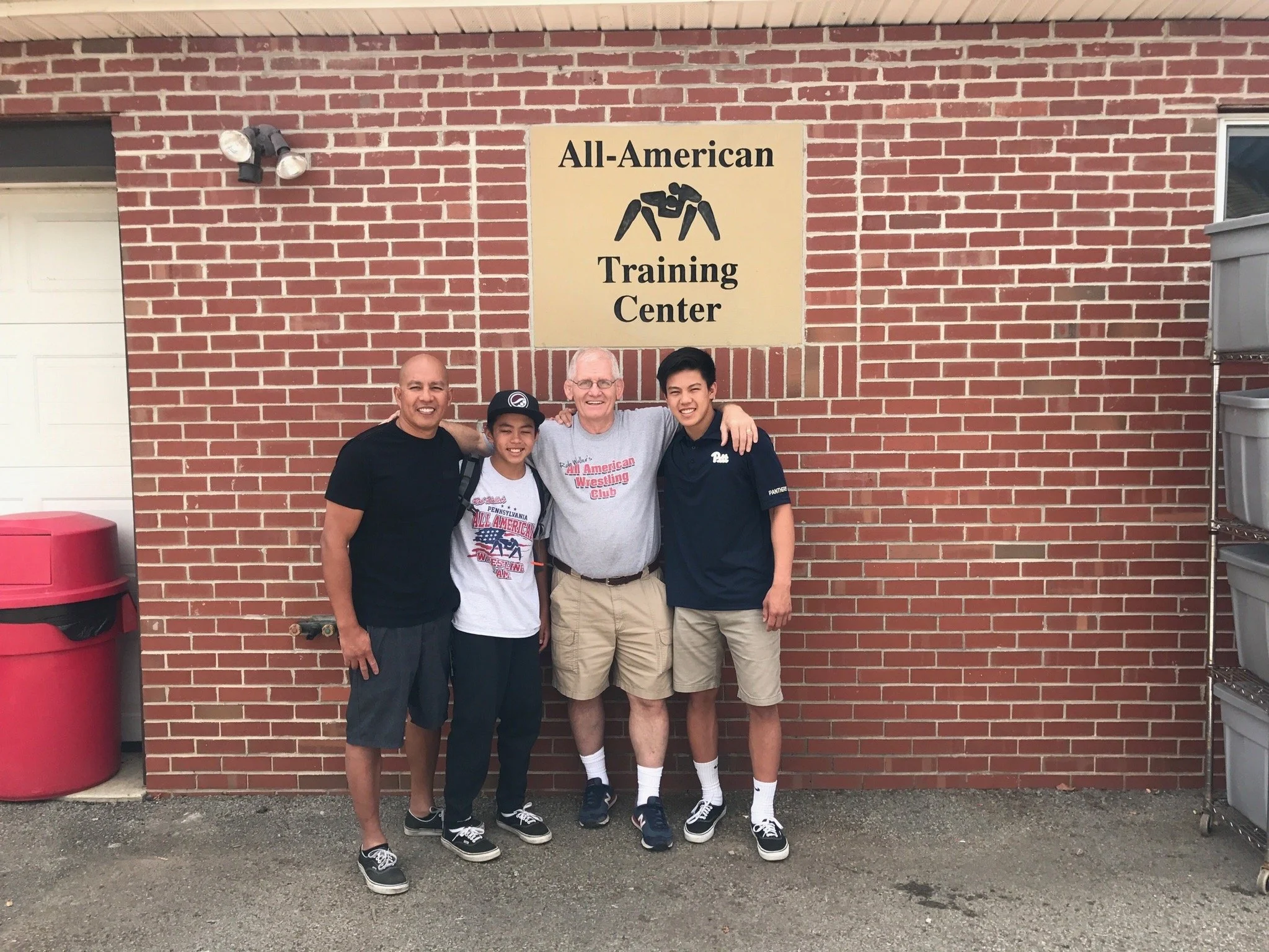 Four people standing in front of a brick wall with a sign that reads 'All-American Training Center,' with three young men and one older man.