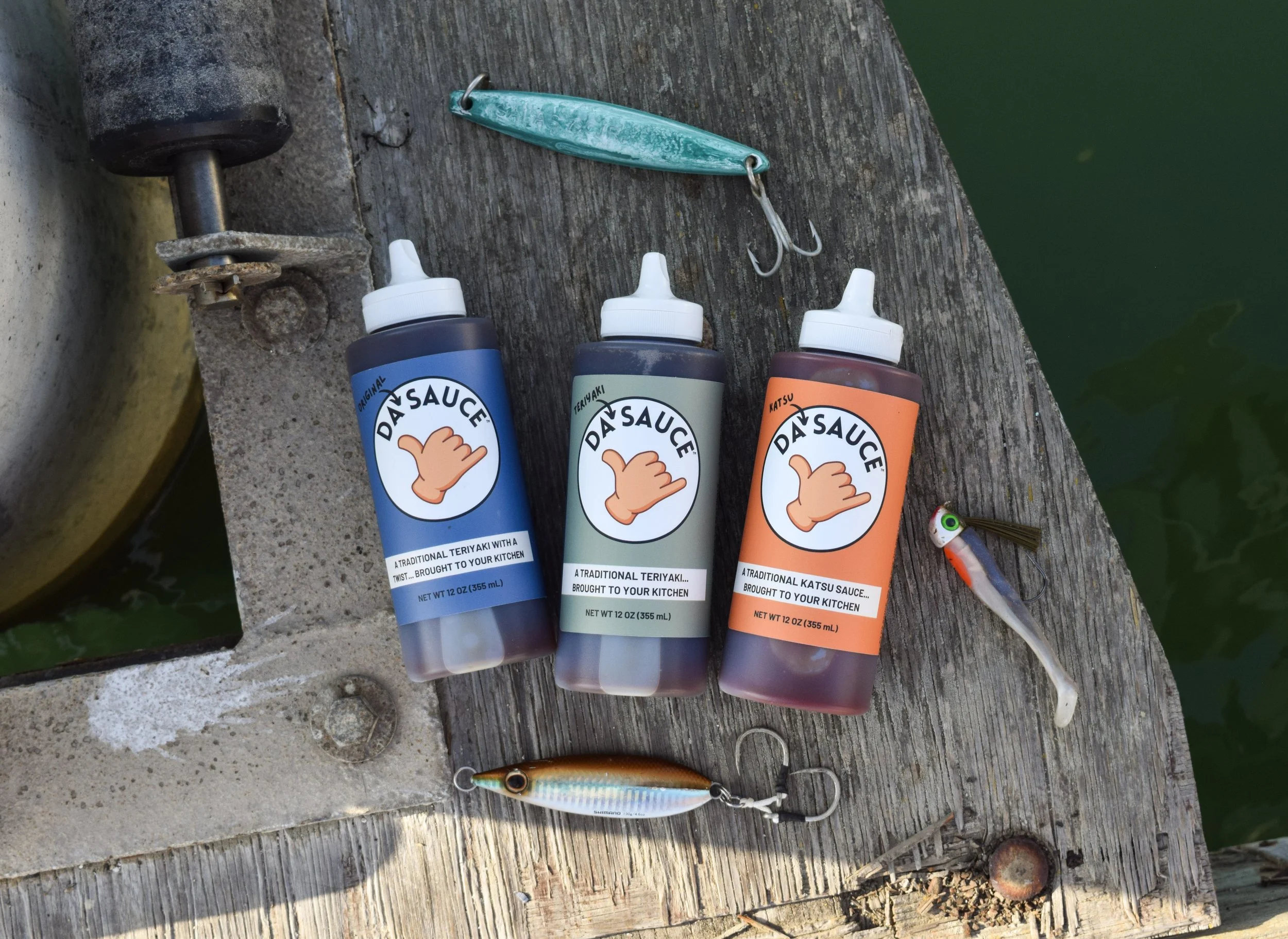 Fishing lures and bottles of sauce on a weathered wooden dock near green water, with a fishing rod tip and part of a fish lure visible.