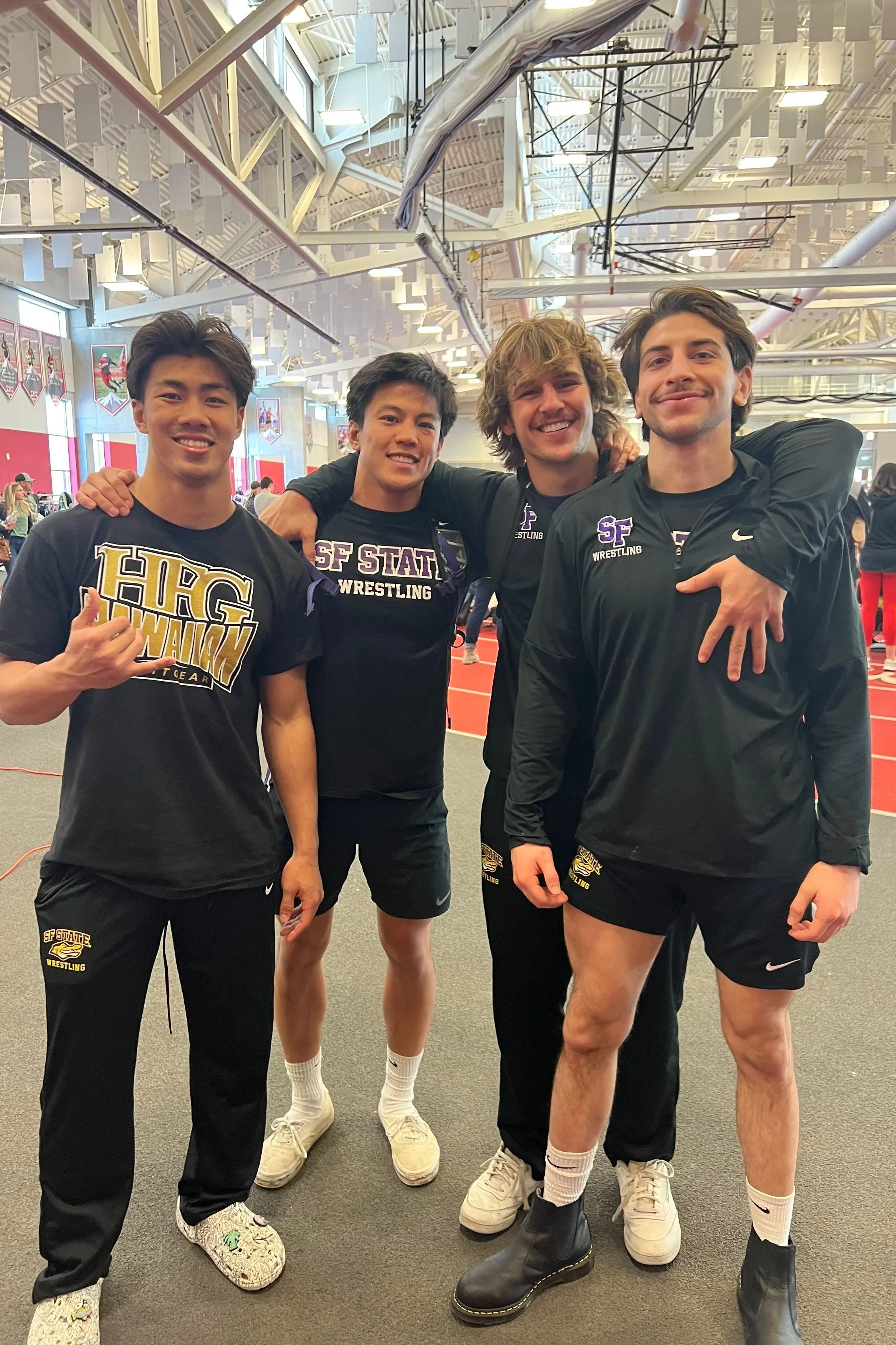 Four male wrestlers in black athletic gear with 'SF State Wrestling' logos, standing inside a sports facility, smiling and posing for a photo.