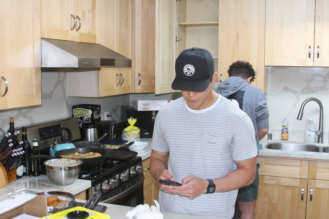 Two young men in a kitchen; one is wearing a black cap and striped t-shirt, looking at a smartphone, the other has curly hair, wearing a gray hoodie, facing away near the sink.