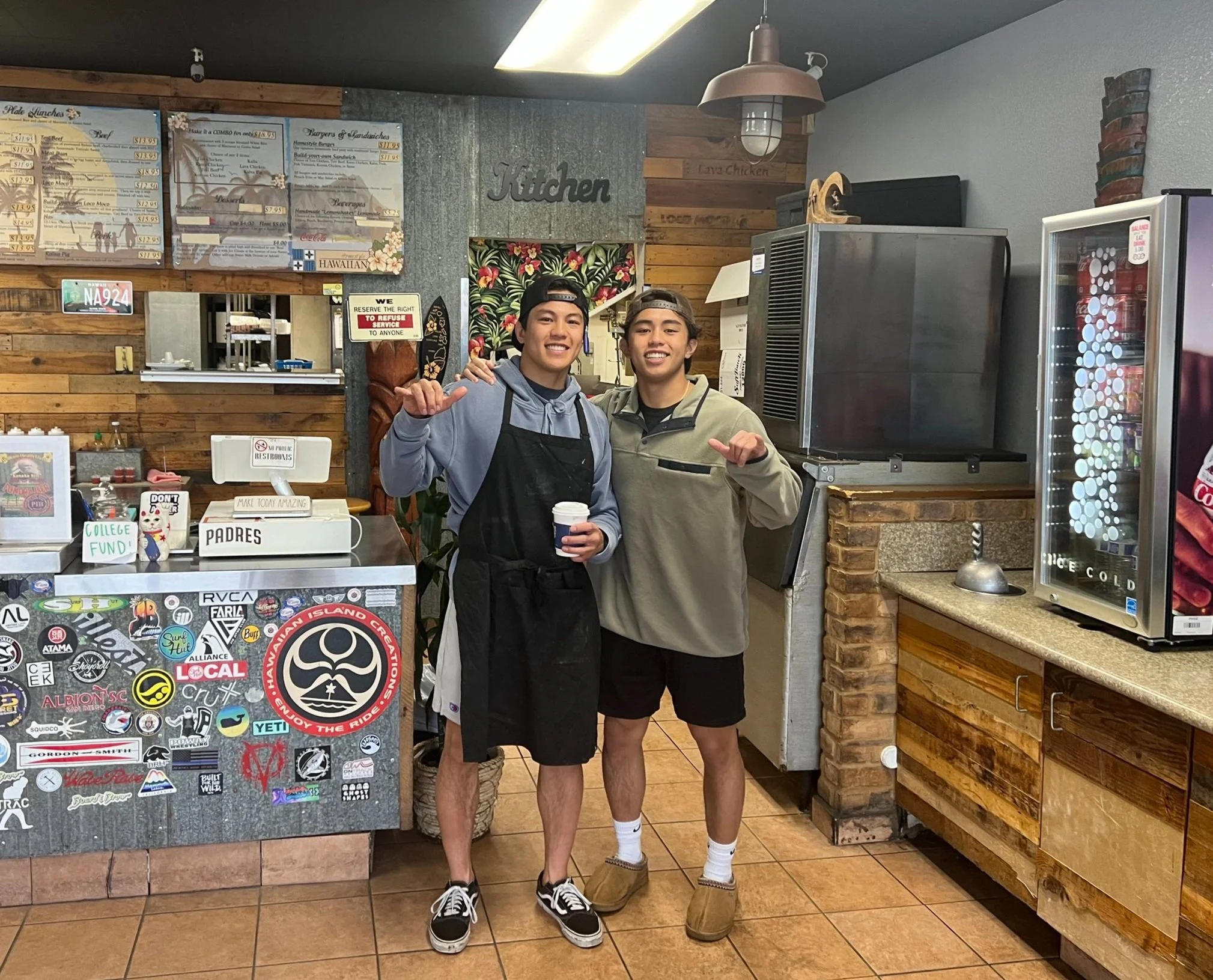 Two young men standing inside a restaurant behind the counter, smiling at the camera. One is holding a coffee cup, both making a shaka hand gesture. They are wearing casual clothing and hats. The background includes menus, a sign with the restaurant's name, and a display fridge.