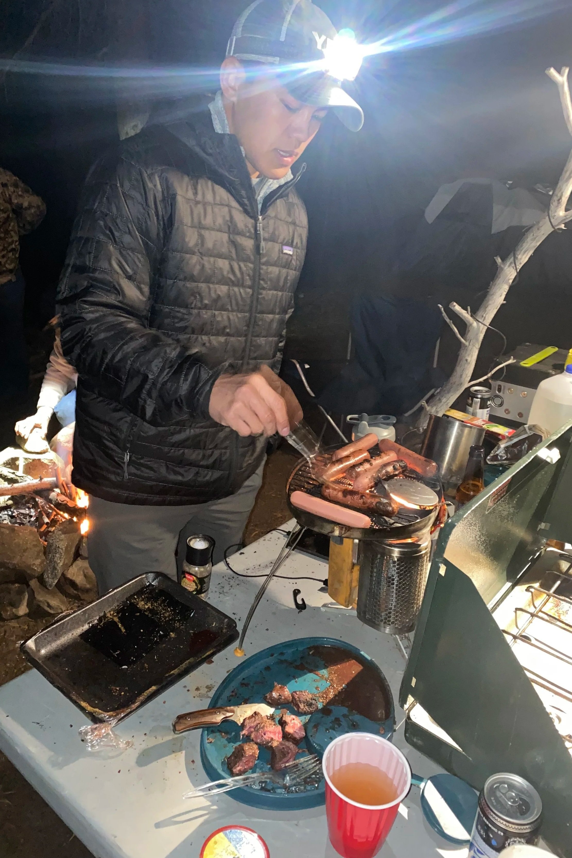 Person cooking hot dogs and meat over a grill at an outdoor campsite at night with camping gear and cooking supplies on a table.