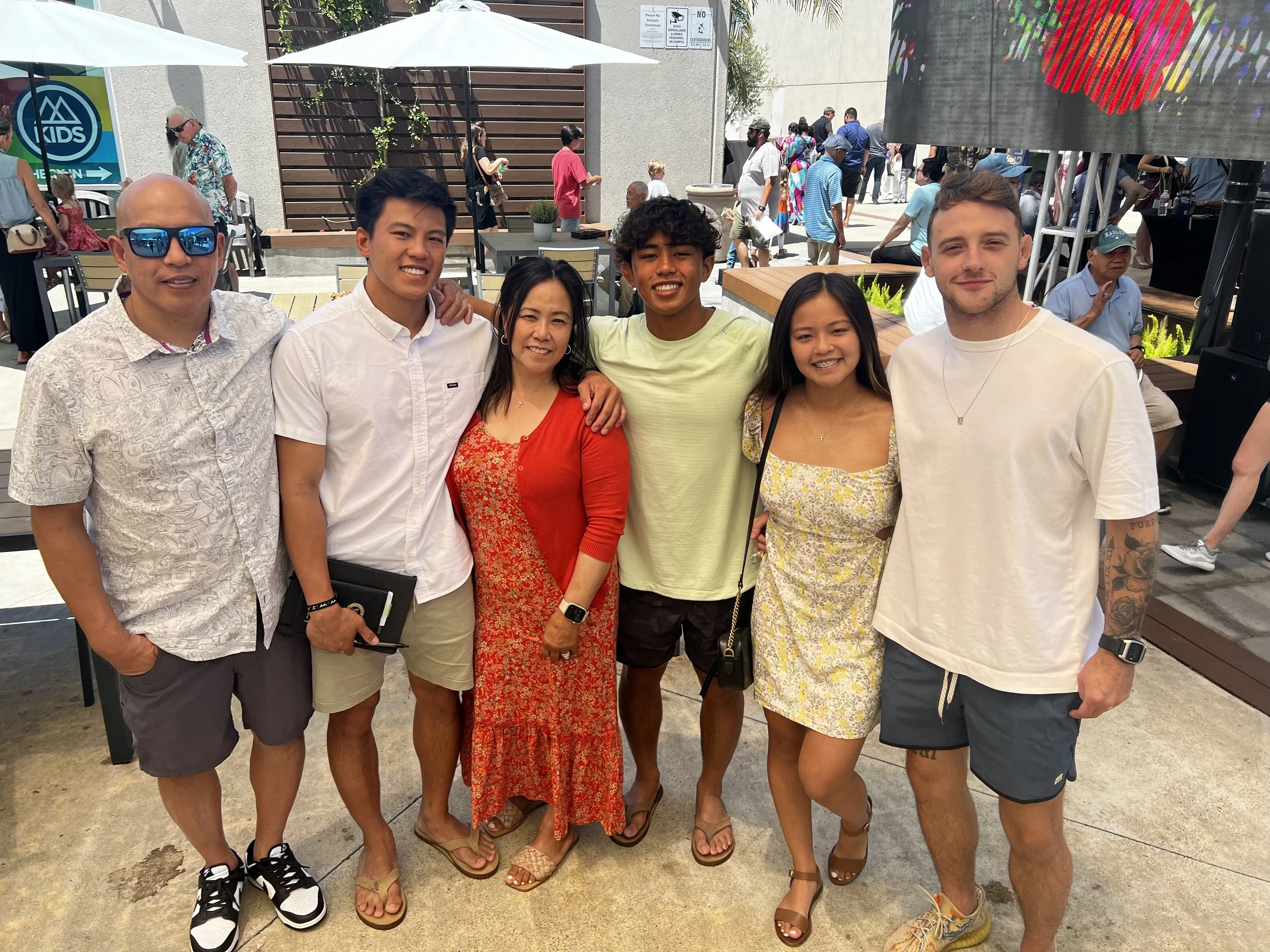 Group of six people standing close together outdoors, smiling at the camera, with other people in the background at a public event. The group includes four men and two women, dressed casually in summer clothing.