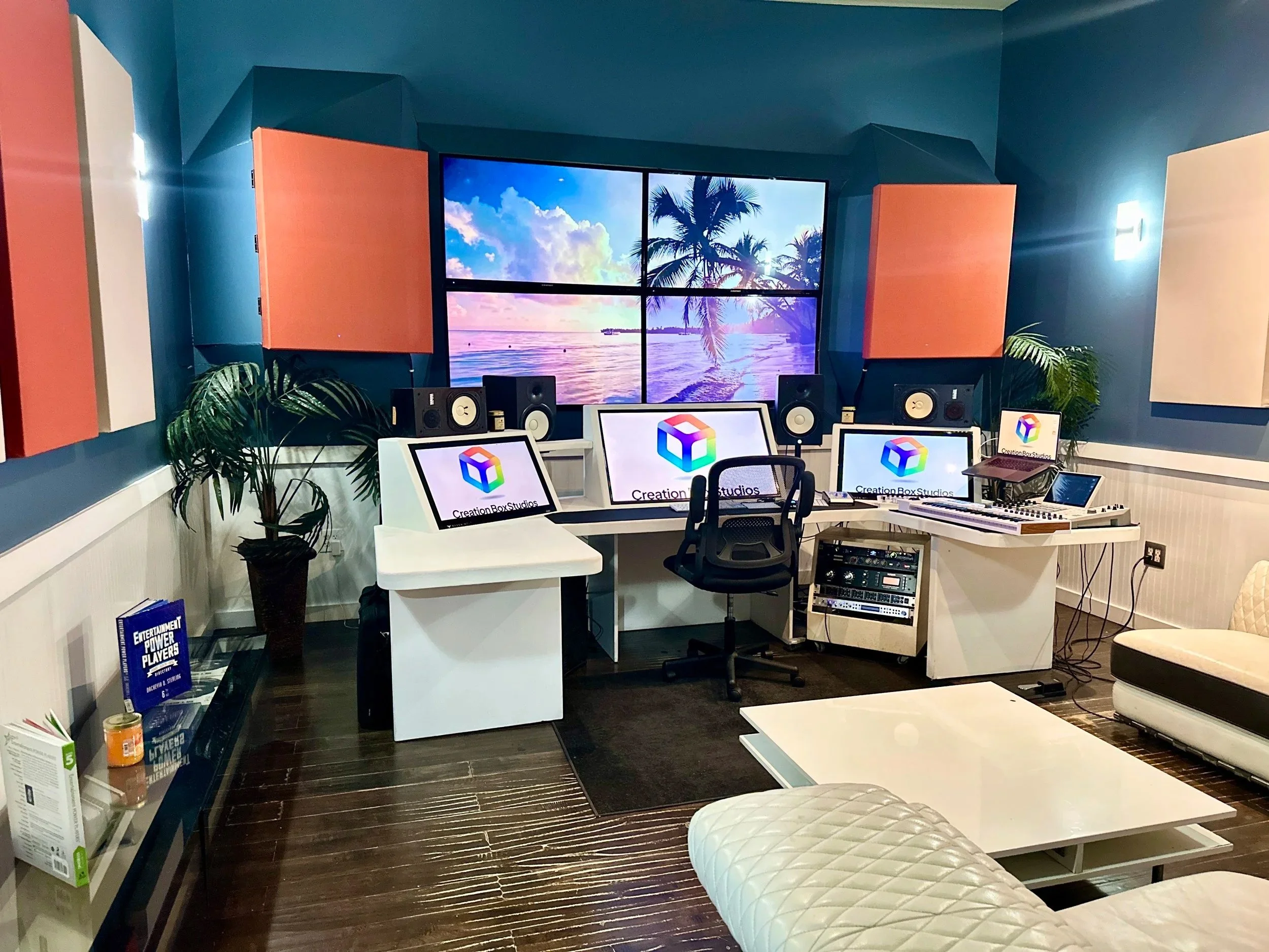 A music production studio with a large wall-mounted display showing a beach scene with palm trees and ocean, multiple computer monitors and studio speakers on a curved white desk, acoustic panels on the teal-colored walls, and a seating area with a white quilted ottoman and a beige chair.