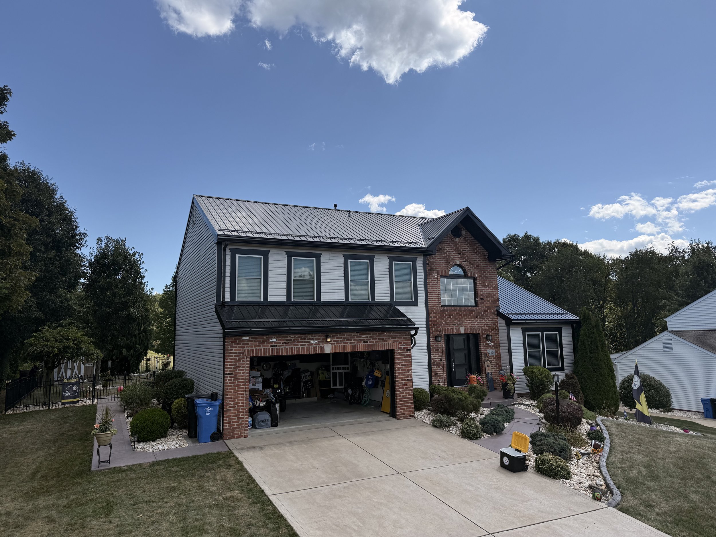 A two-story house with a brick and white siding exterior, metal roof, and front yard decorated with plants, rocks, and yard toys.
