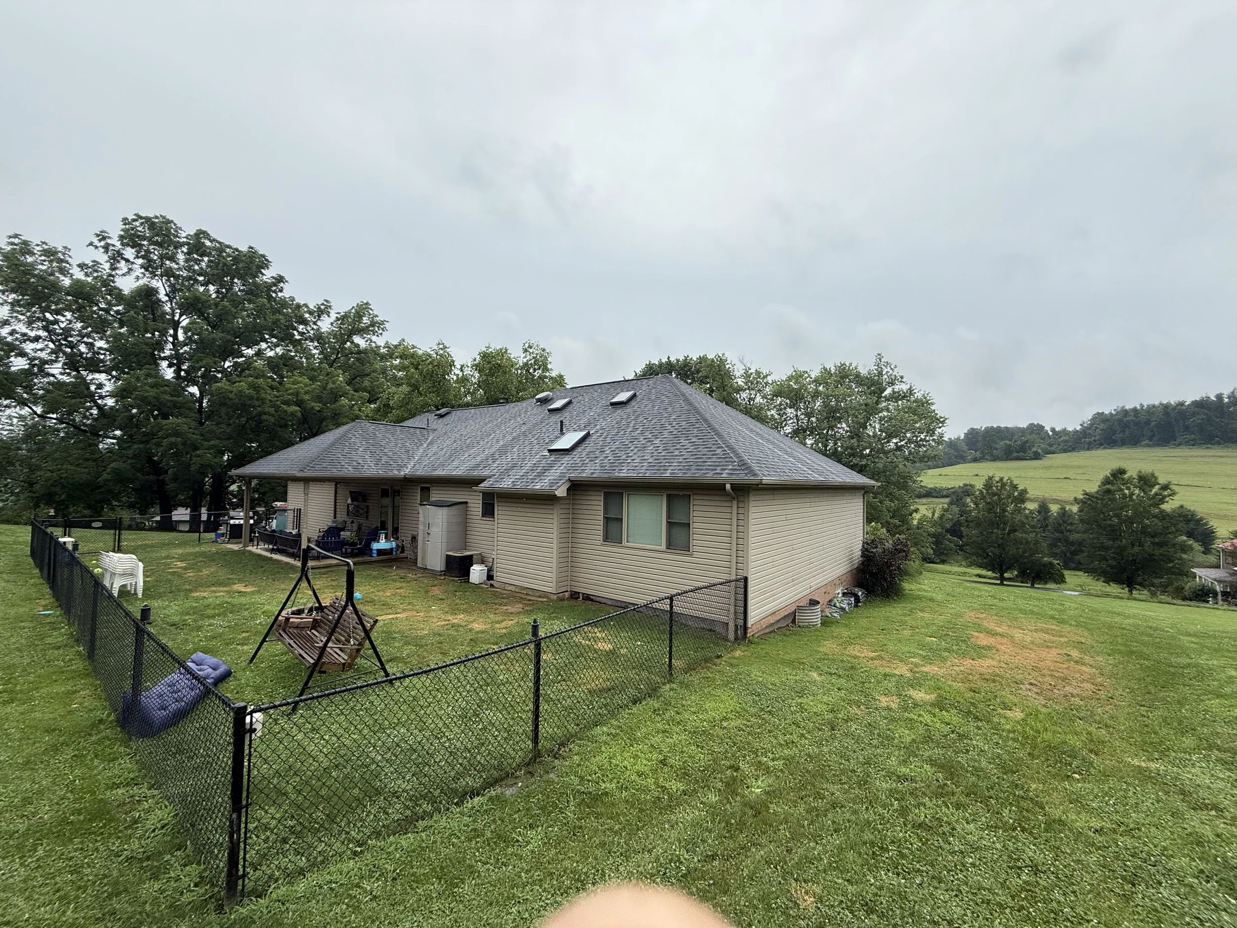 Backyard of a house with a fenced yard, outdoor furniture, and a swing set on a cloudy day, with trees and rolling hills in the background.