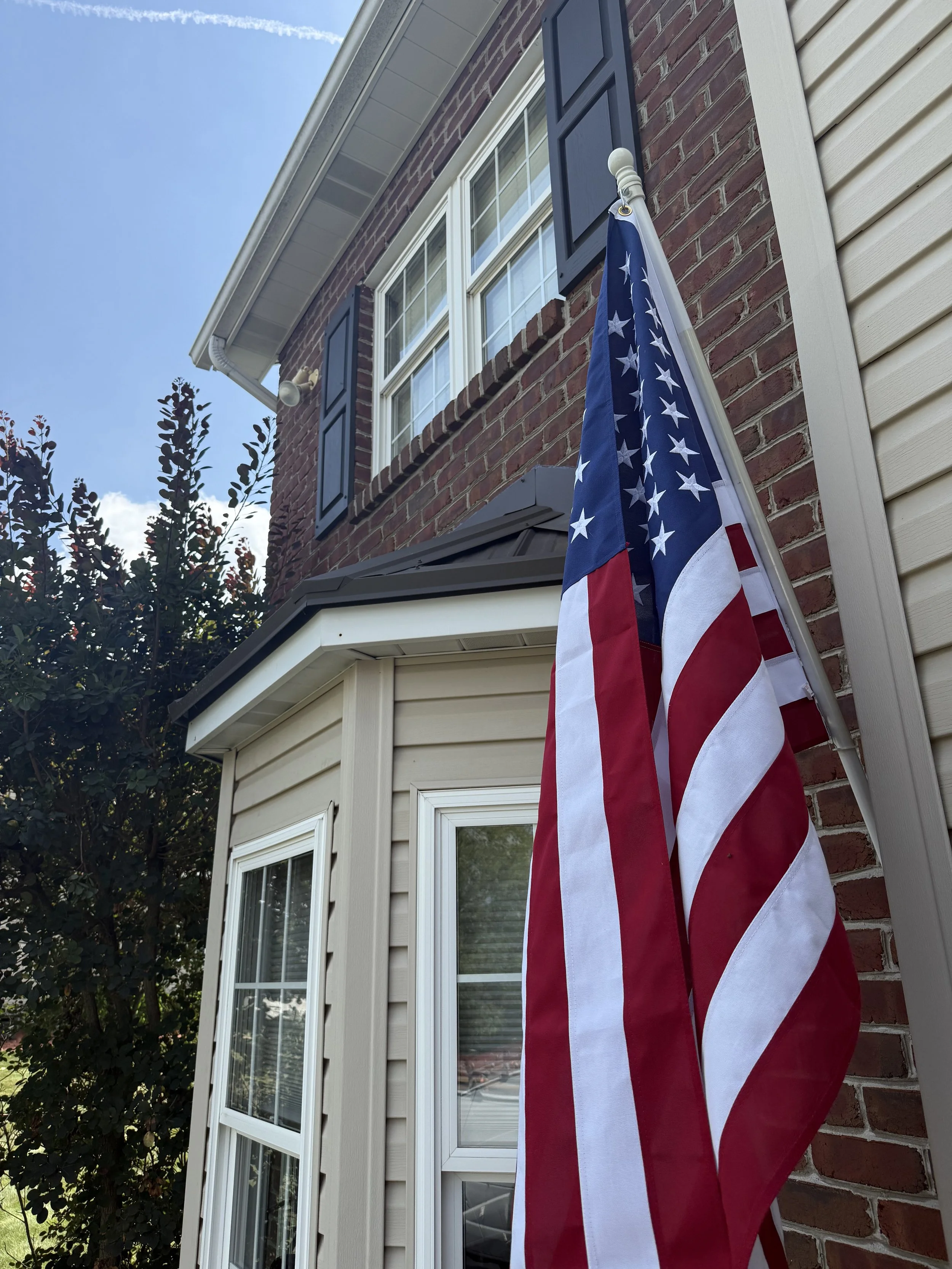 American flag hanging outside a house with beige siding and brick wall, next to a window with white framing and blue shutters.