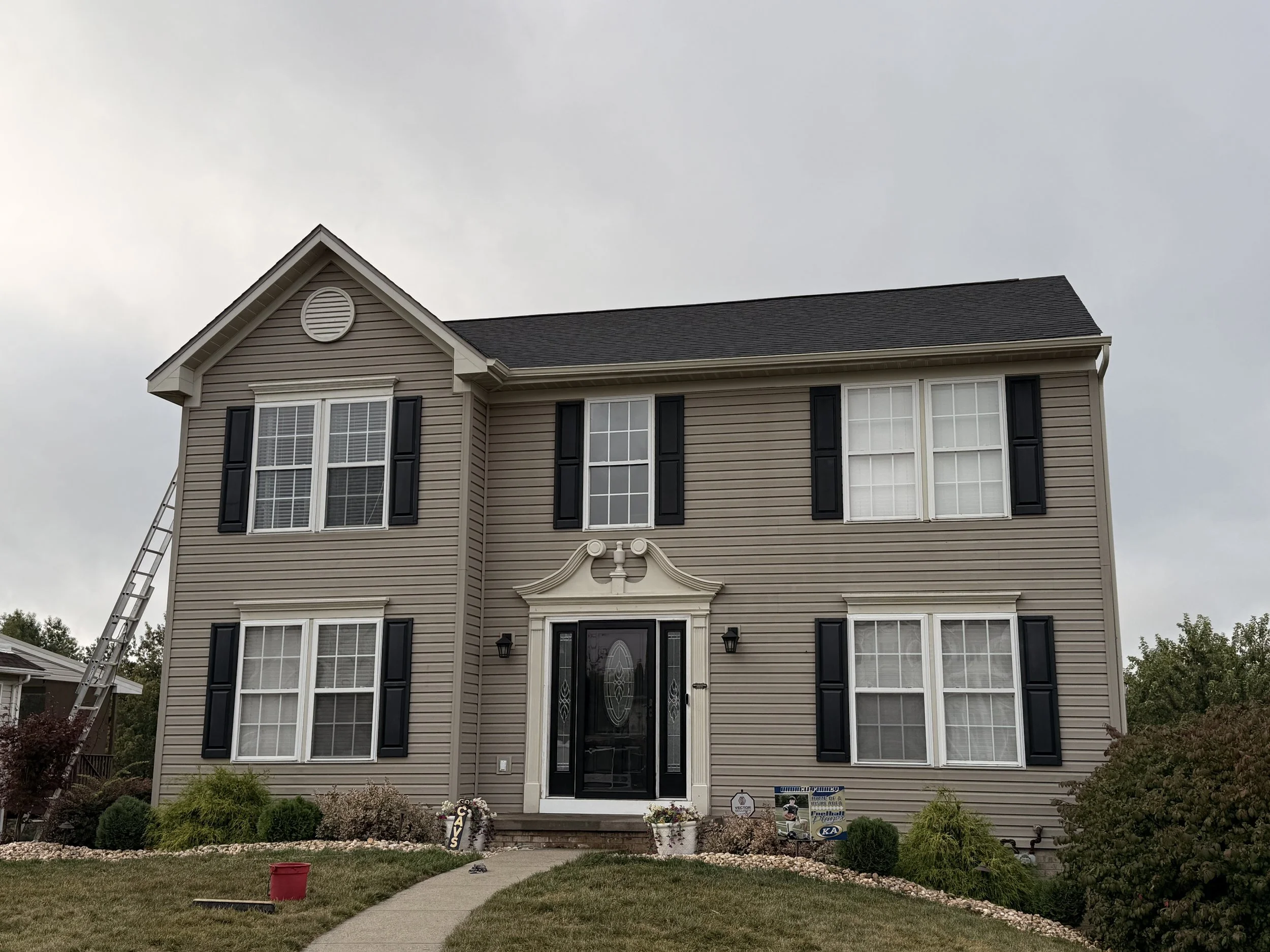 A two-story house with beige siding, black shutters, and a black front door with decorative glass. The house has four window units with white frames, two on each floor. There is landscaping with bushes and a curved walkway leading to the front door, and a ladder leaning against the house on the left side.