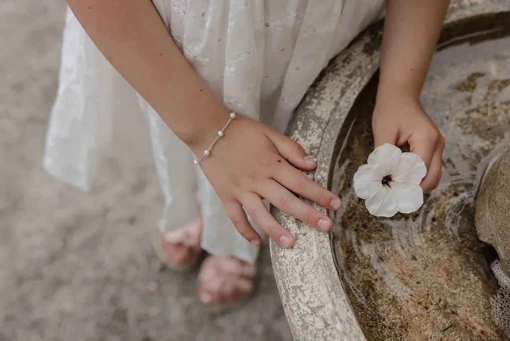 A young girl is holding a flower standing next to a fountain, on her wrist is a pearl station bracelet in sterling silver from Brilliant Fine+Custom jewelry in Roswell, Georgia.