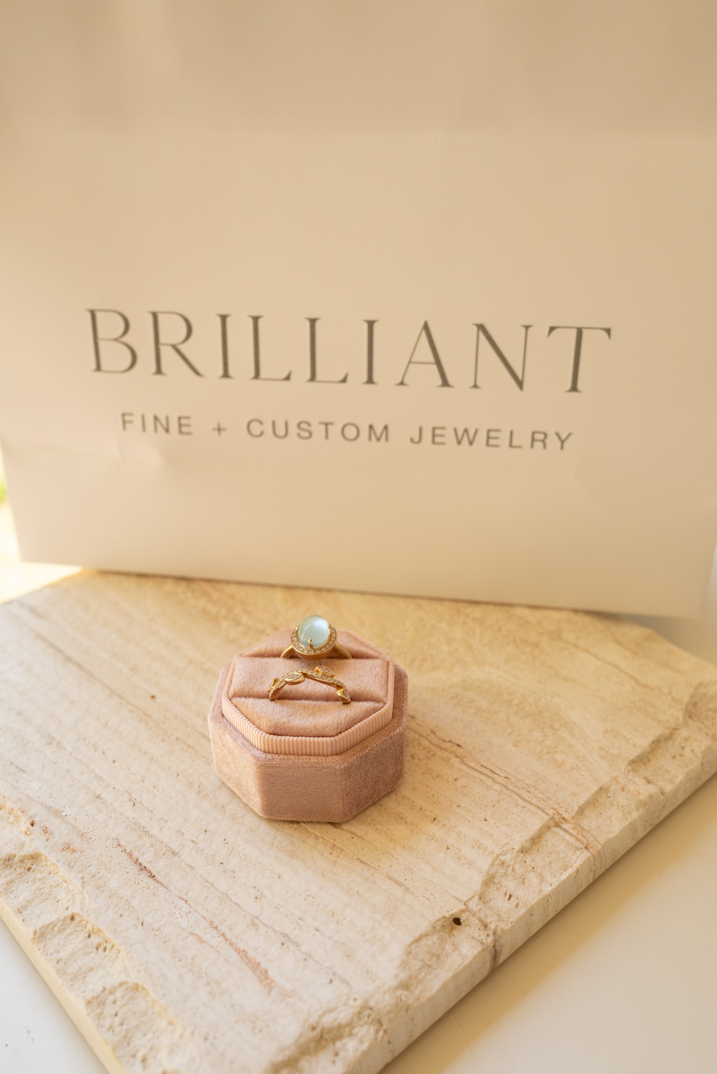 Two rings, part of a ring stack, sitting in an octagonal velvet ring box, in front of a bag which reads "Brilliant Fine+Custom Jewelry"