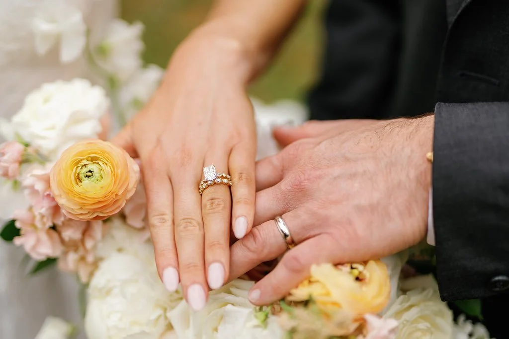 A bride wearing a custom ring stack from Brilliant Fine+Custom jewelry places her hand on top of her husband's hand atop floral decorations.