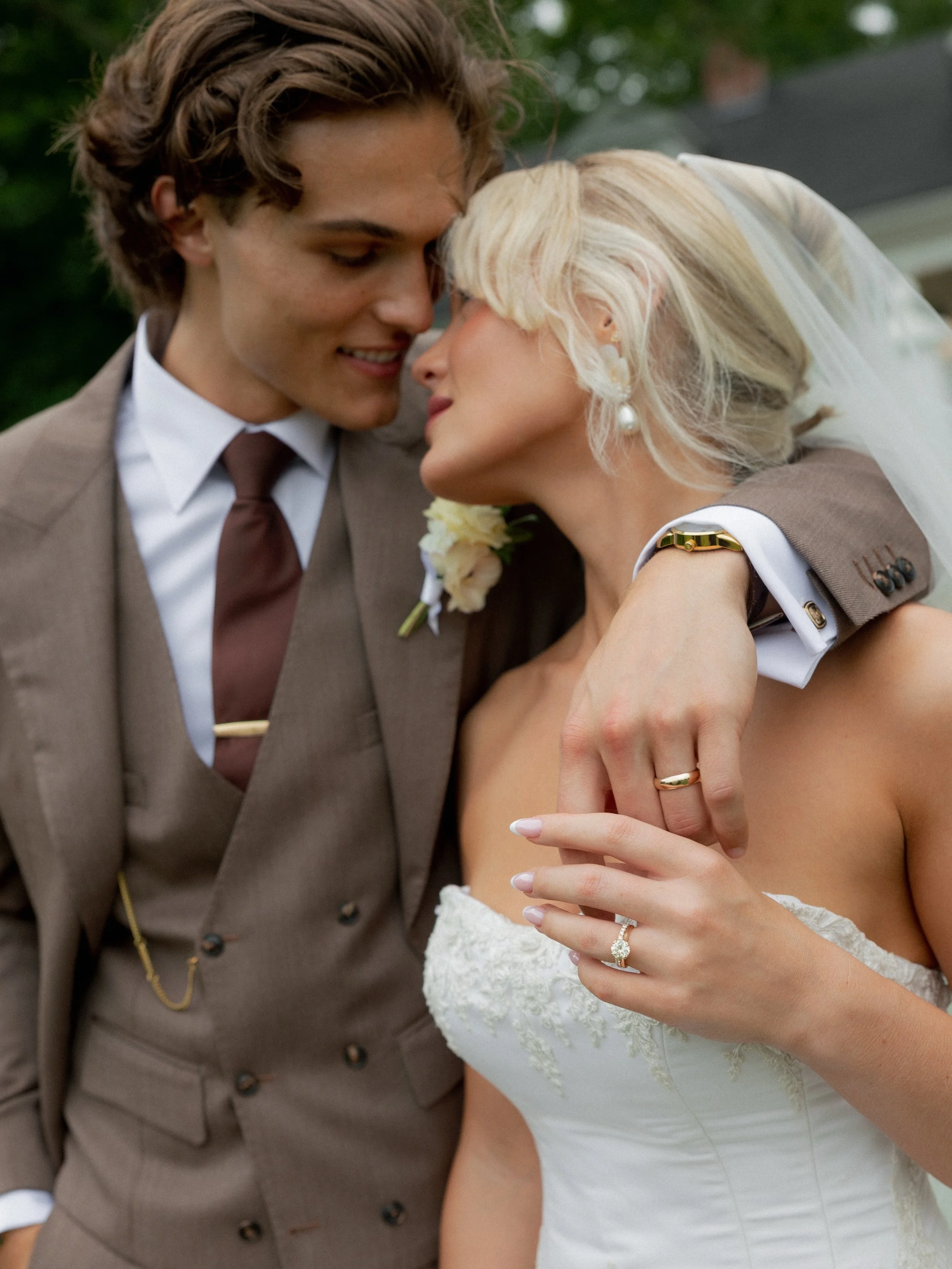 A Bride and Groom embrace and nuzzle each other while  showing the bride's wedding ring and the groom's wedding band.