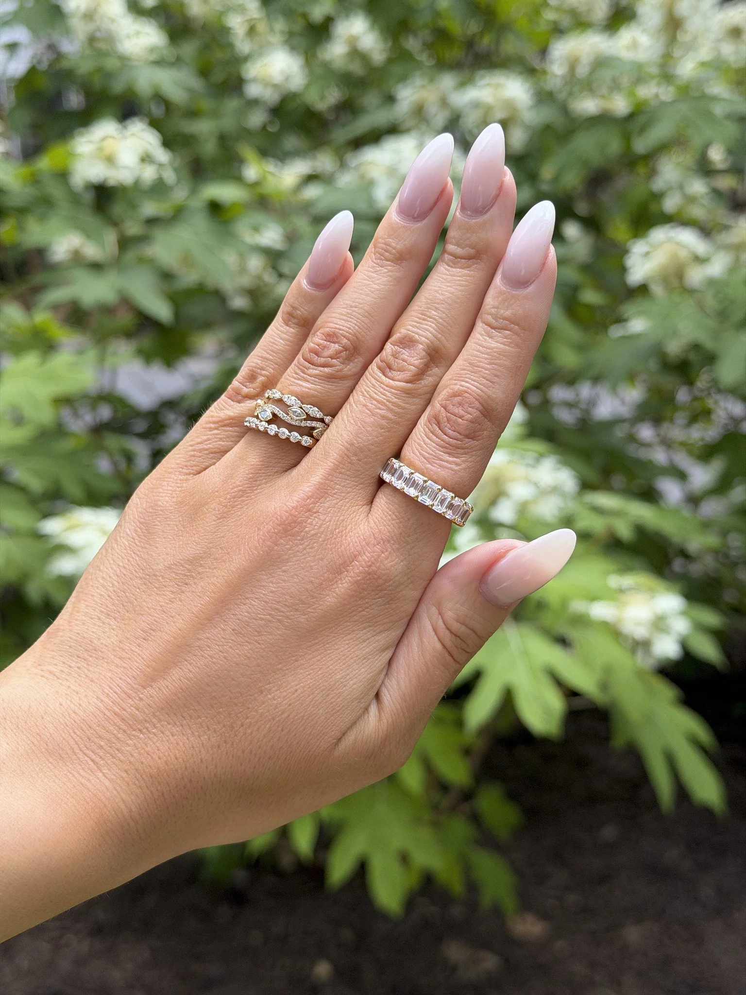 A lady's hand showing off a ring stack on her ring finger with an elegant combination of different diamond rings set in white gold. On her index finger she's wearing a band with multiple emerald-cut stones, all by Brilliant Fine+Custom Jewelry.