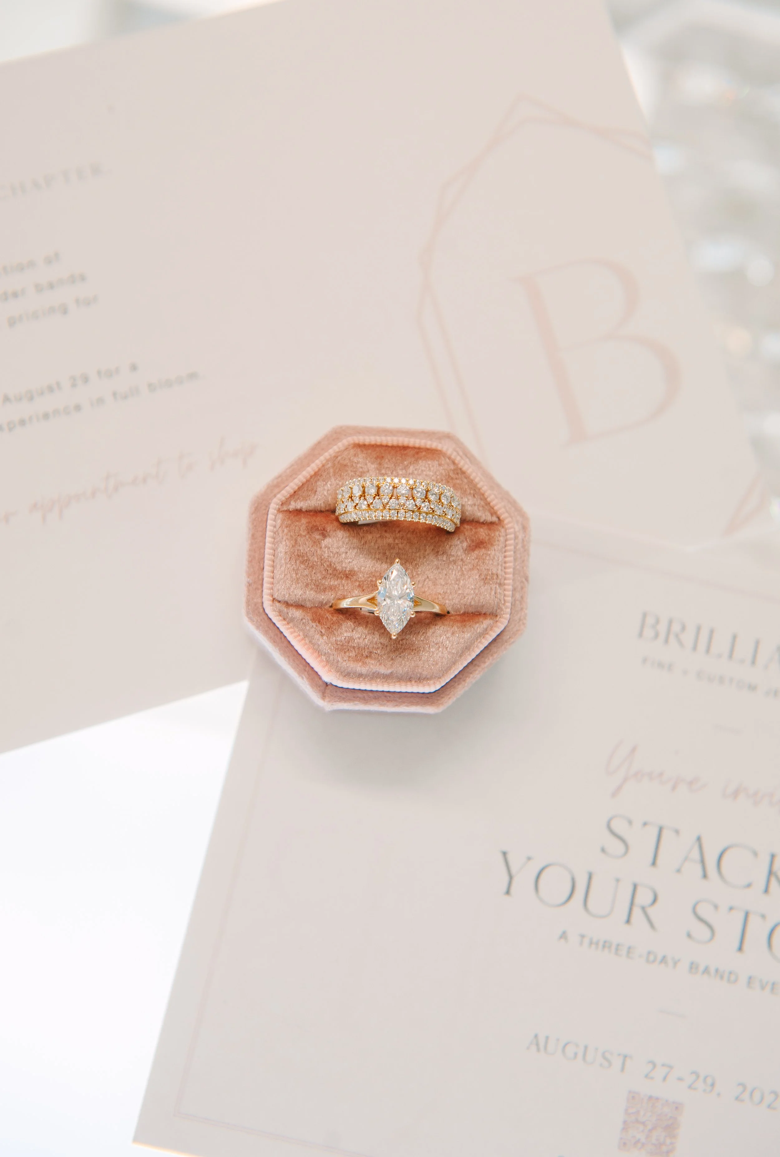 Marquise diamond engagement ring and stacked diamond bands in a blush velvet ring box beside the Stack Your Story event invitation.