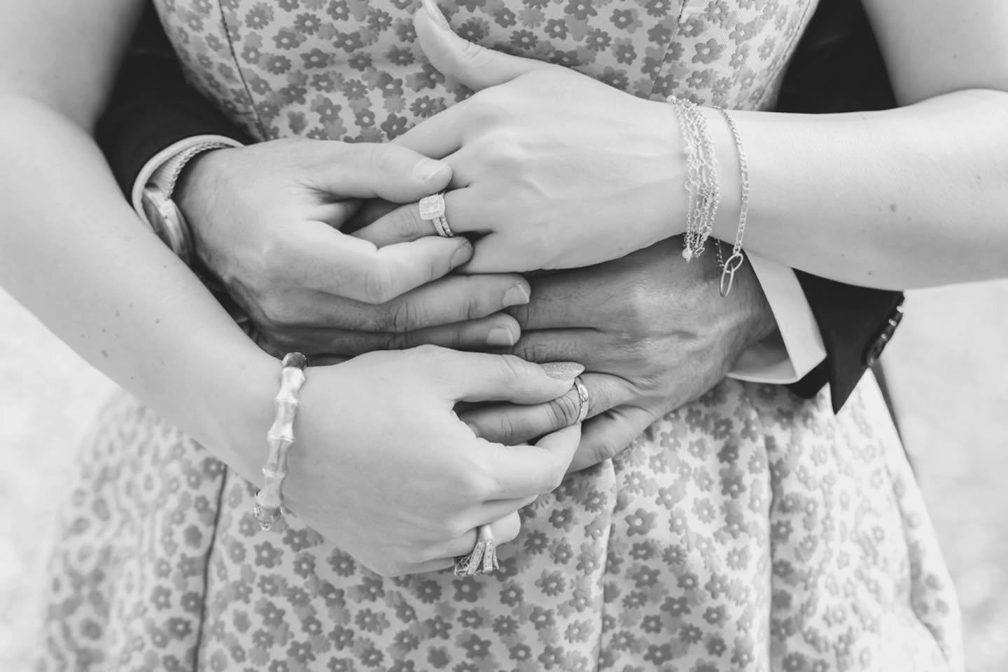 A black and white image of a married couple, with the bride's ring stack of her wedding and engagement rings on prominent display as she locks hands with her husband who is wearing a solid gold wedding band.