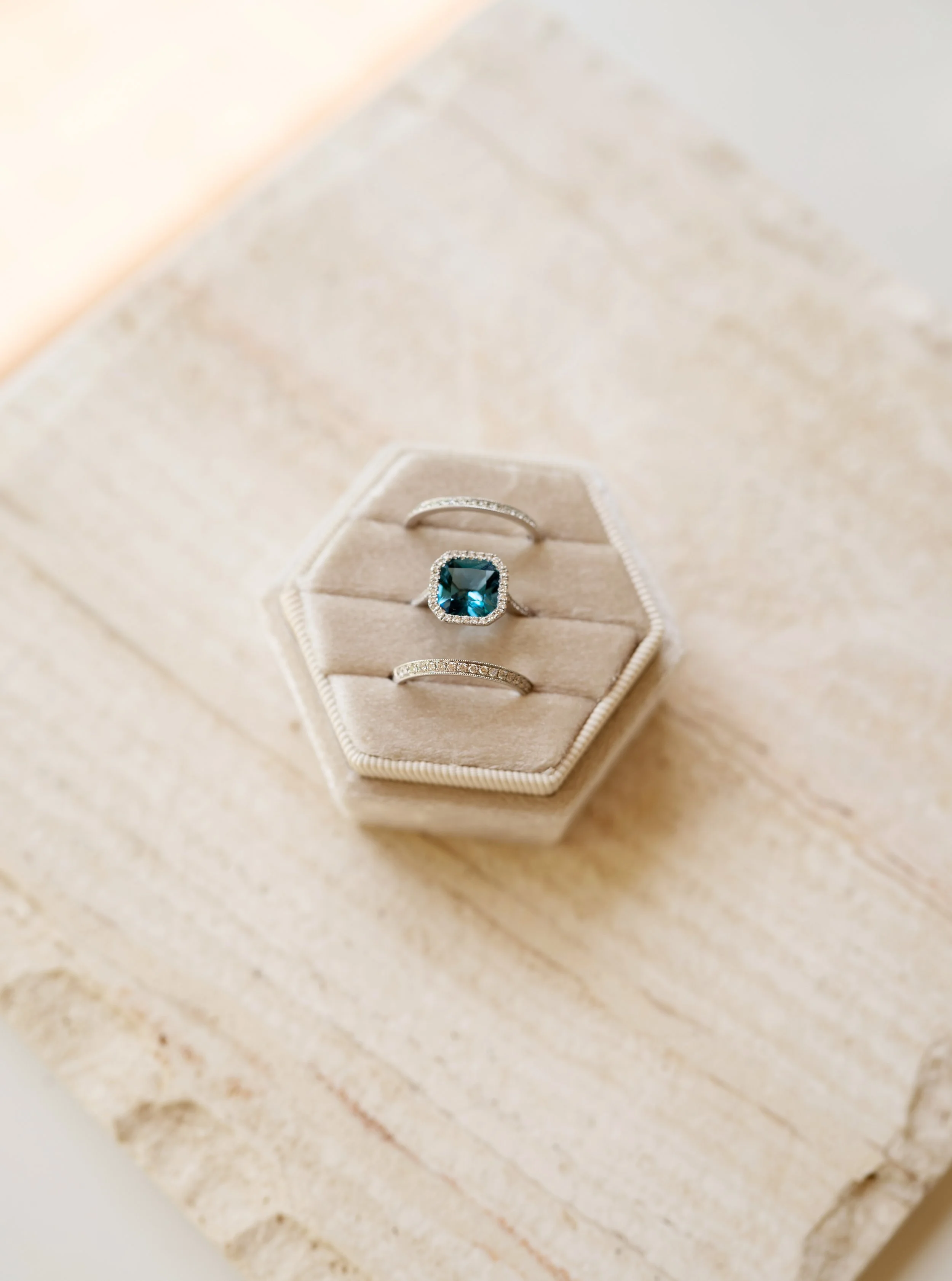 Three stackable rings in a velvet ring box. The top and bottom rings are Eternity Bands with diamonds embedded into the band. The center ring is a emerald-square cut Sapphire stone with diamond halo. All rings are from Brilliant Fine+Custom Jewelry.